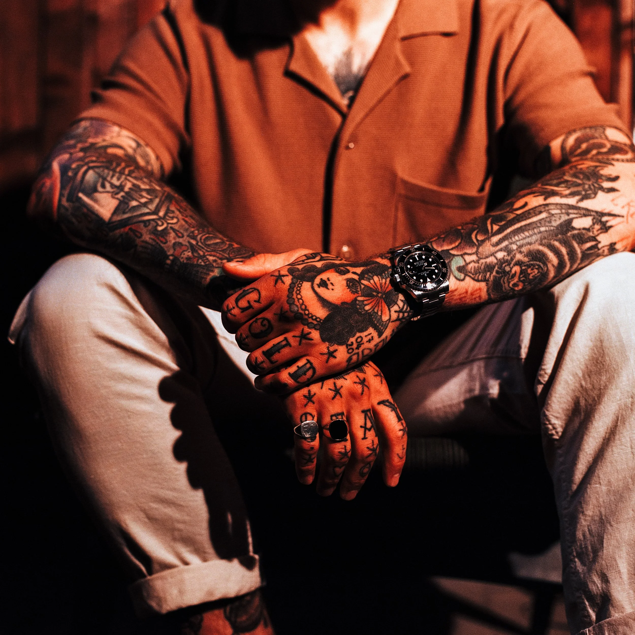A person with heavily tattooed arms and hands, wearing a tan short-sleeve shirt and light-colored pants, sits with legs apart. They are wearing a black wristwatch and several rings, with a red lighting setting in the background.