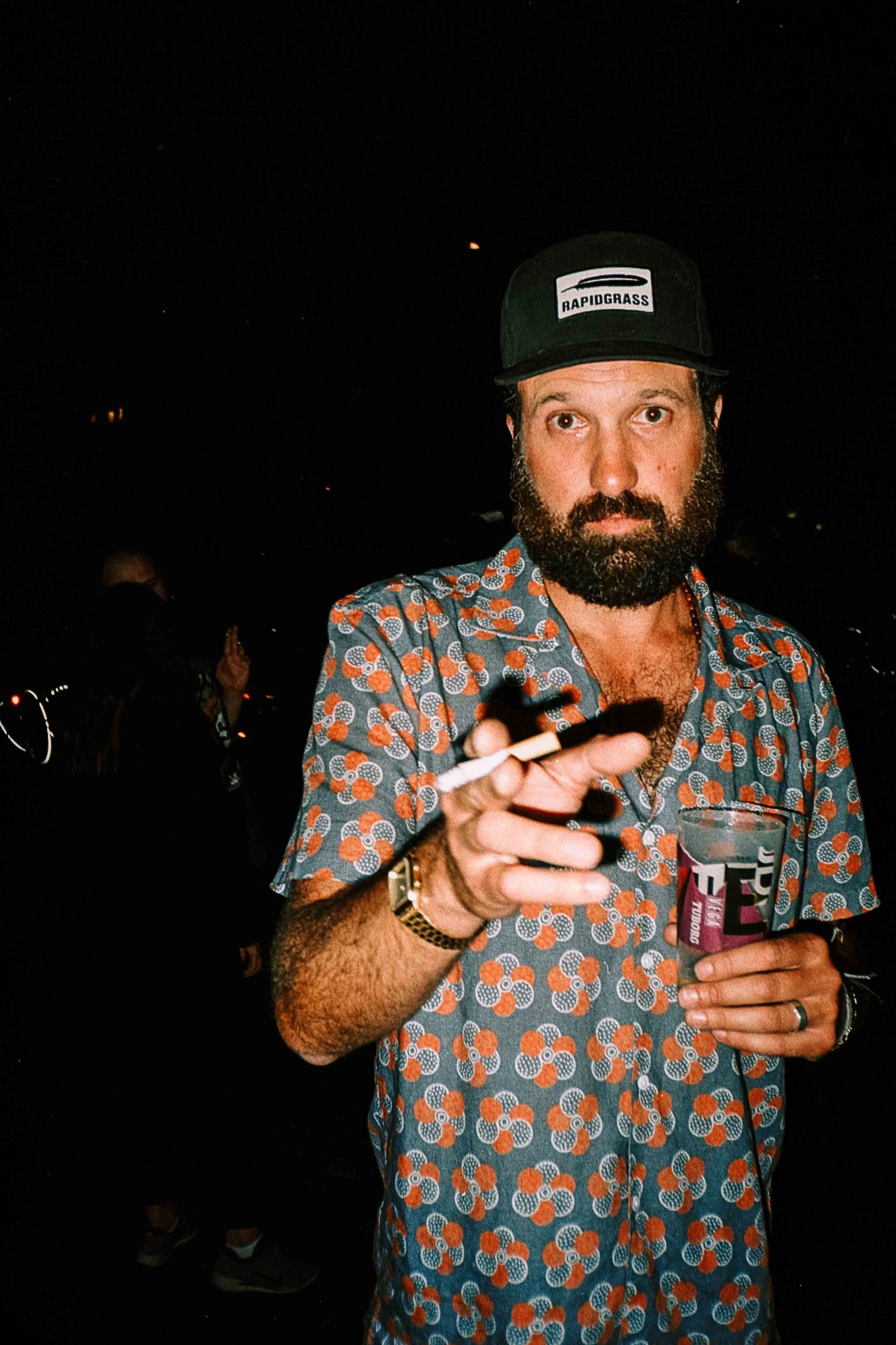 A man with a beard wearing a patterned shirt and a cap labeled 'Rapidgrass,' holding a drink and a cigarette at night.
