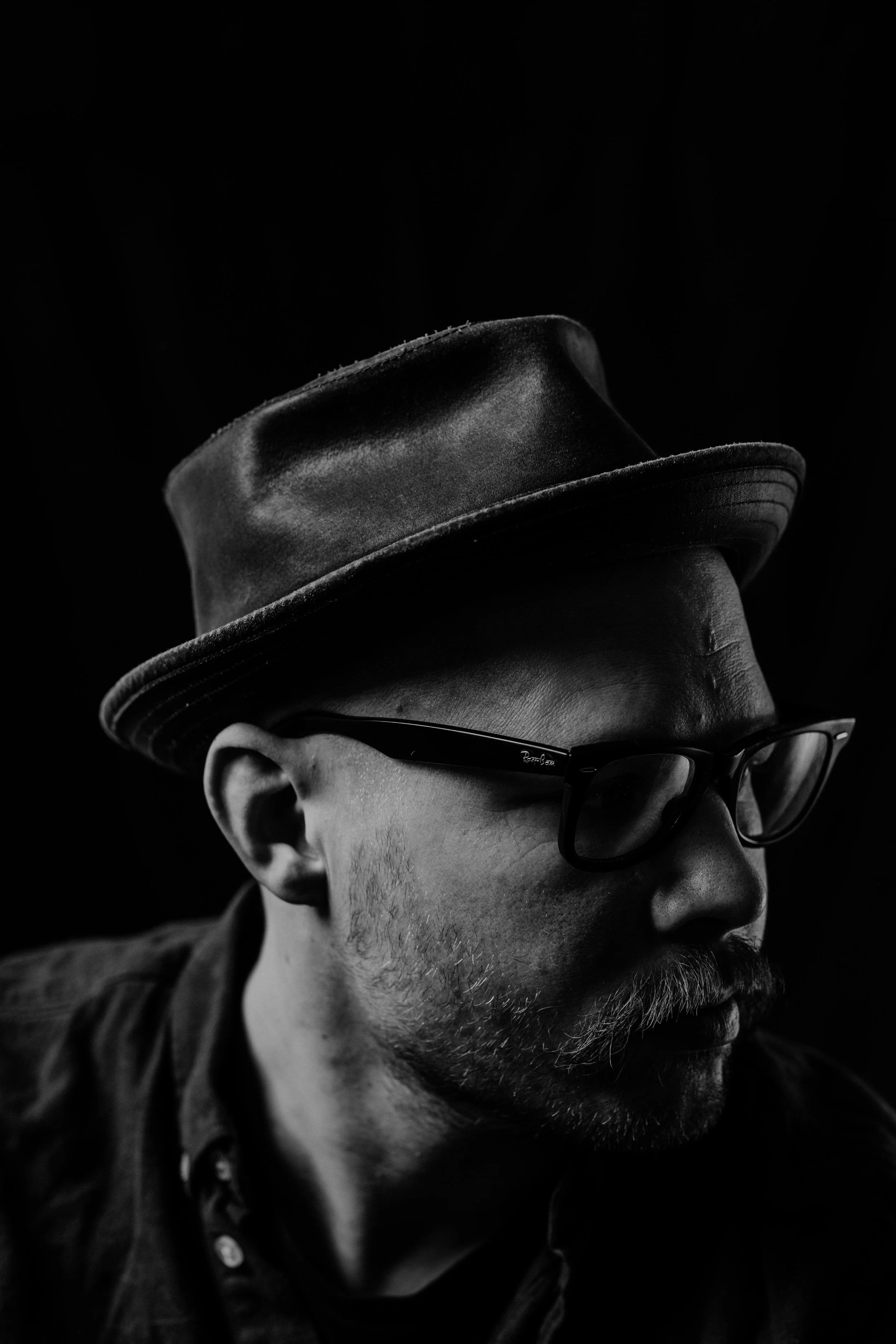 Black and white portrait of a man wearing glasses, a hat, and a jacket, in profile view against a dark background.