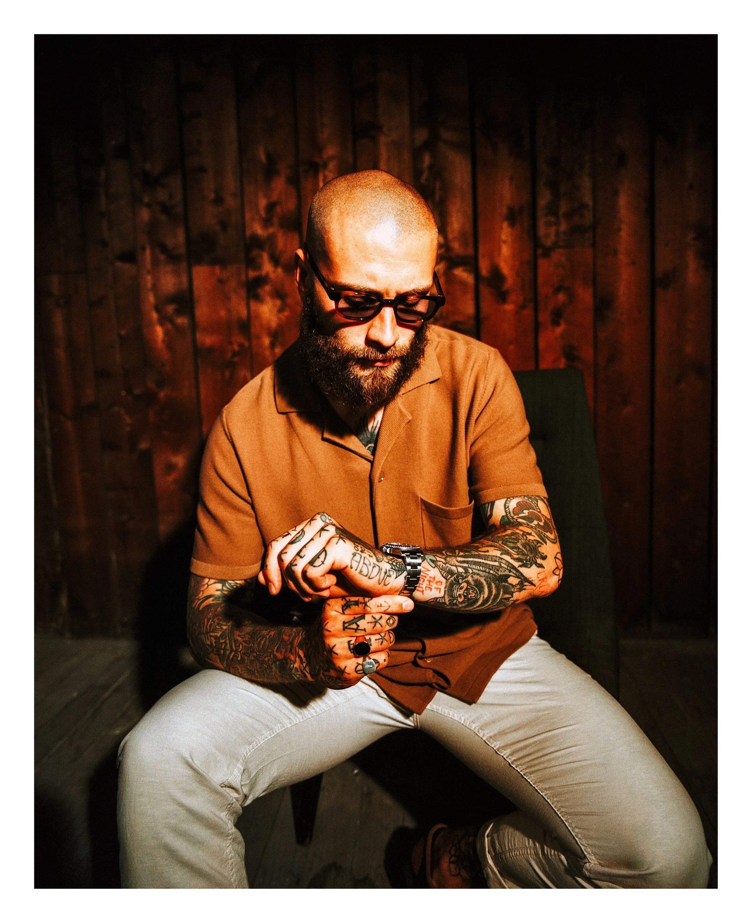 A bearded man with glasses, tattoos, and a watch sits in a wooden-paneled room, looking at his wristwatch.