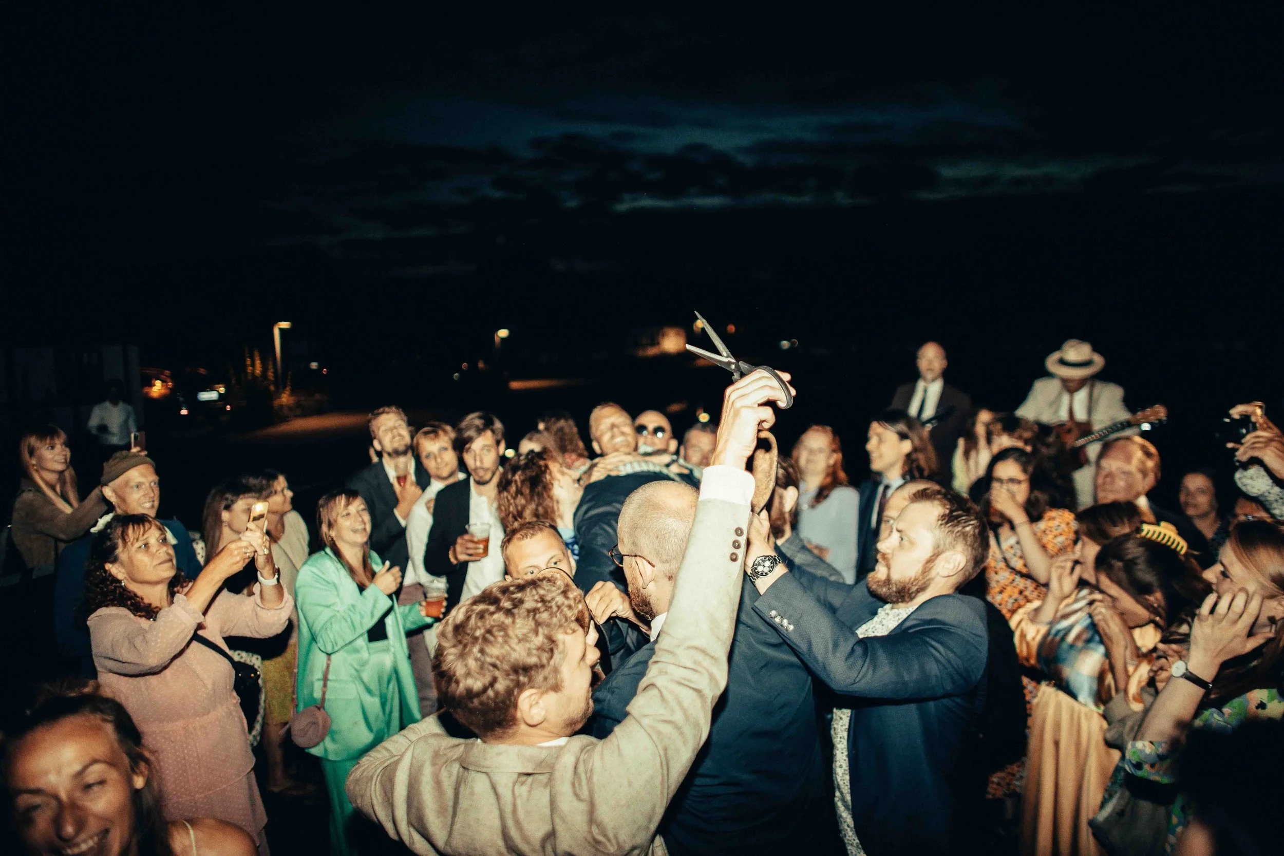 Crowd of people at night, some taking photos, others dancing, with a musician playing guitar in the background.