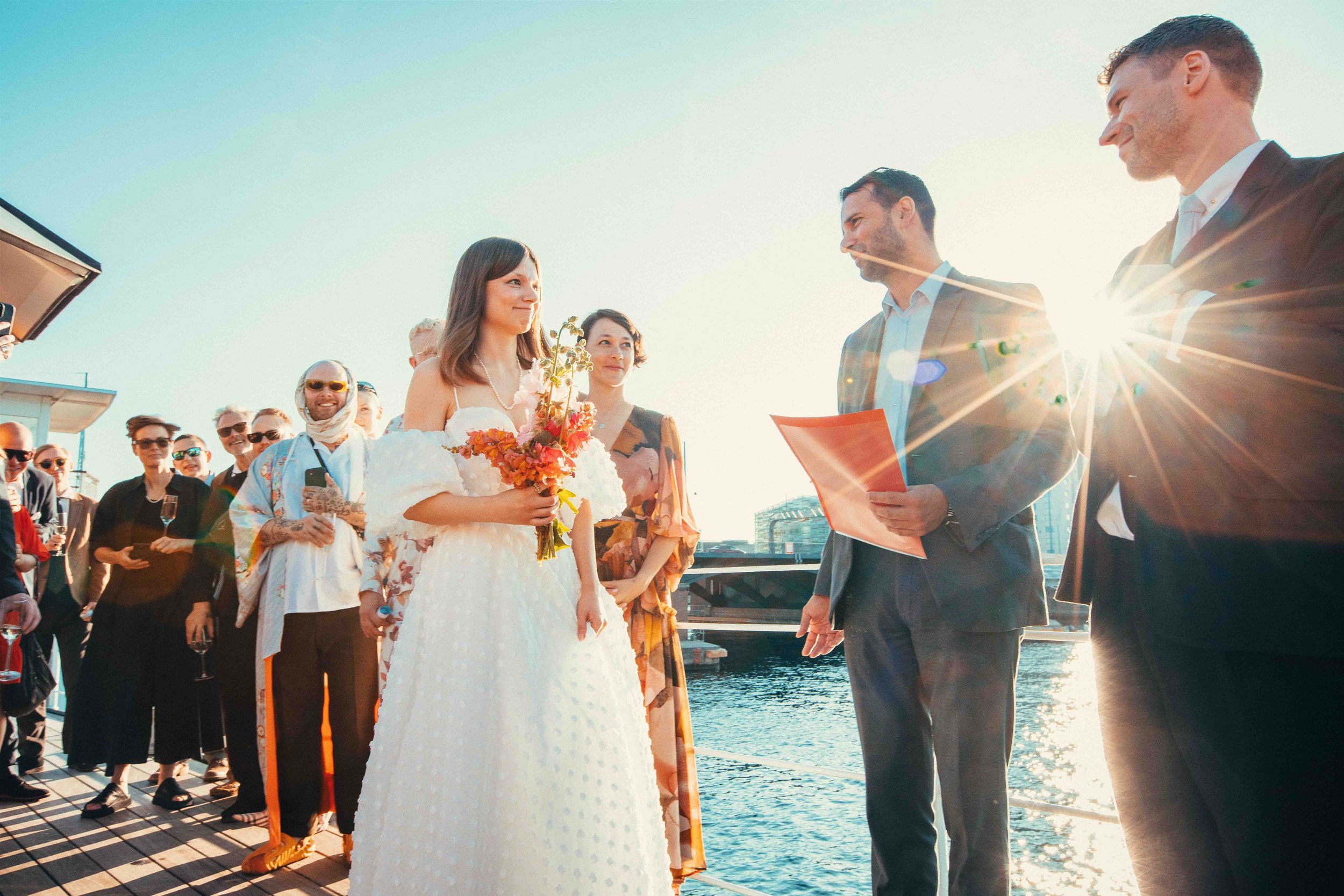A woman in a white wedding dress holding a bouquet of flowers, standing on a dock during a wedding ceremony with friends and family gathered nearby, with water and buildings in the background and sunlight creating a lens flare.
