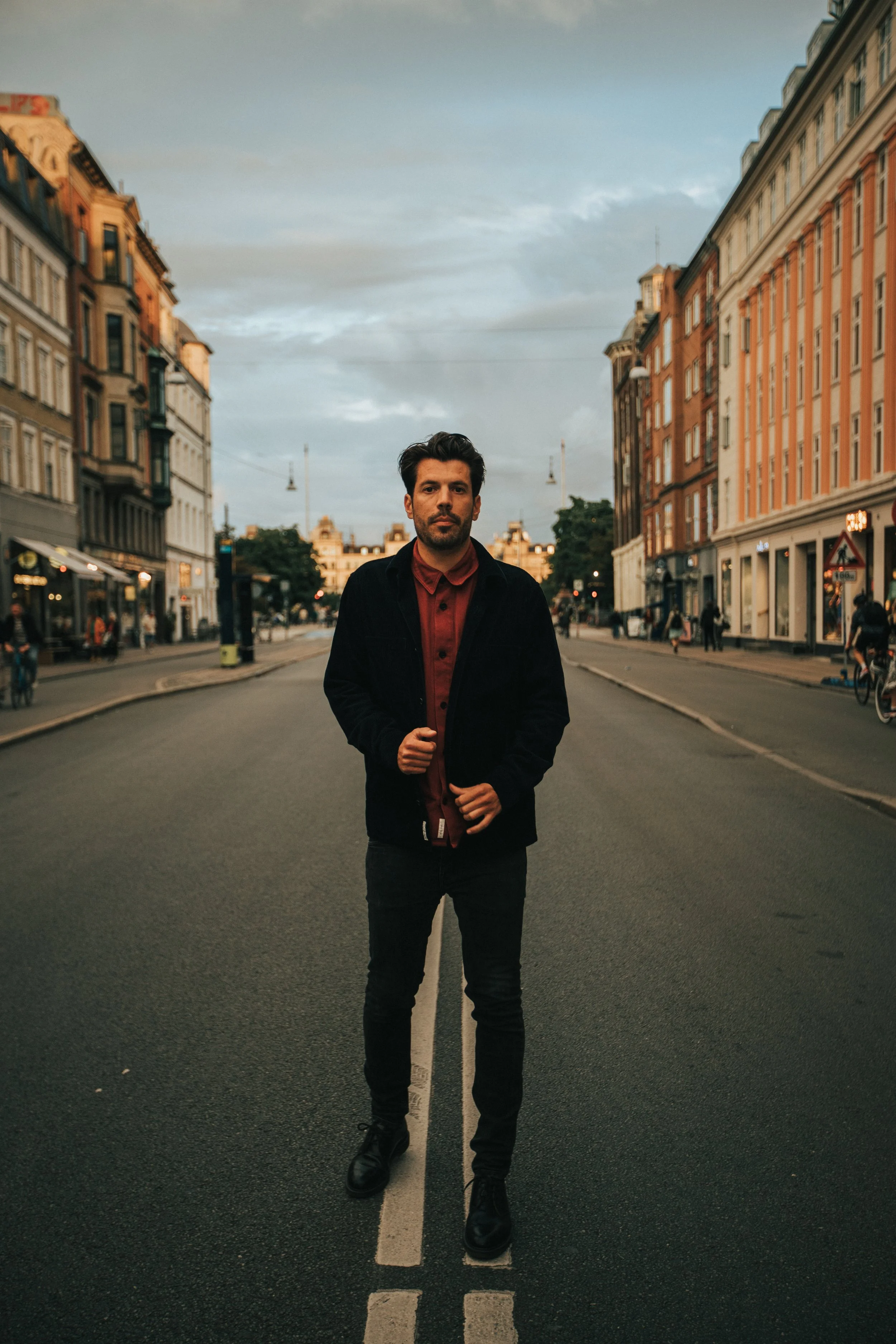 A man is standing in the middle of a city street during dusk, holding the front of his coat with both hands, surrounded by historic buildings on either side.