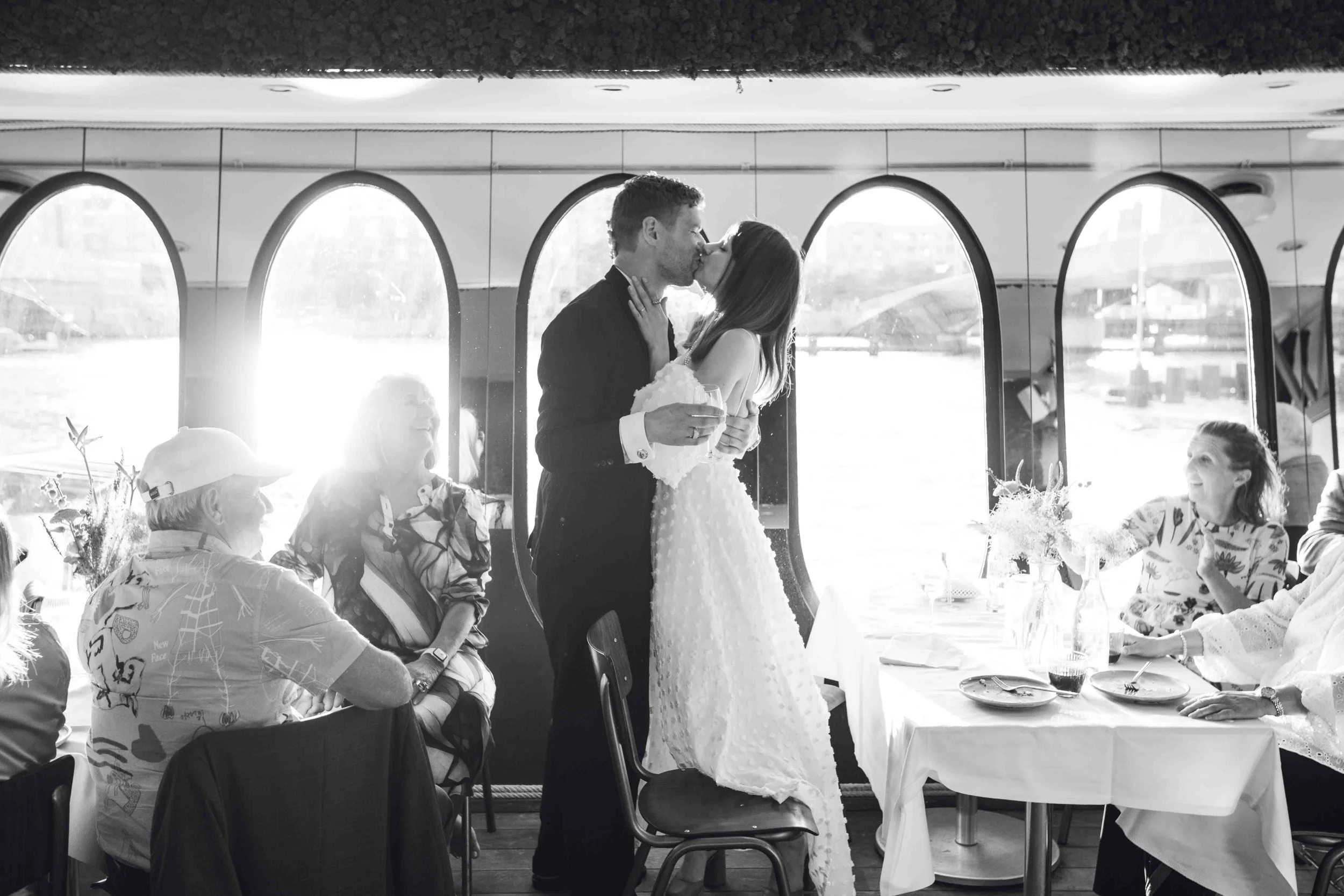 A black-and-white photo of a wedding reception with a bride and groom sharing a kiss inside a room with large arched windows. The bride is wearing a white dress and the groom is in a dark suit. Guests are seated at tables with floral arrangements and