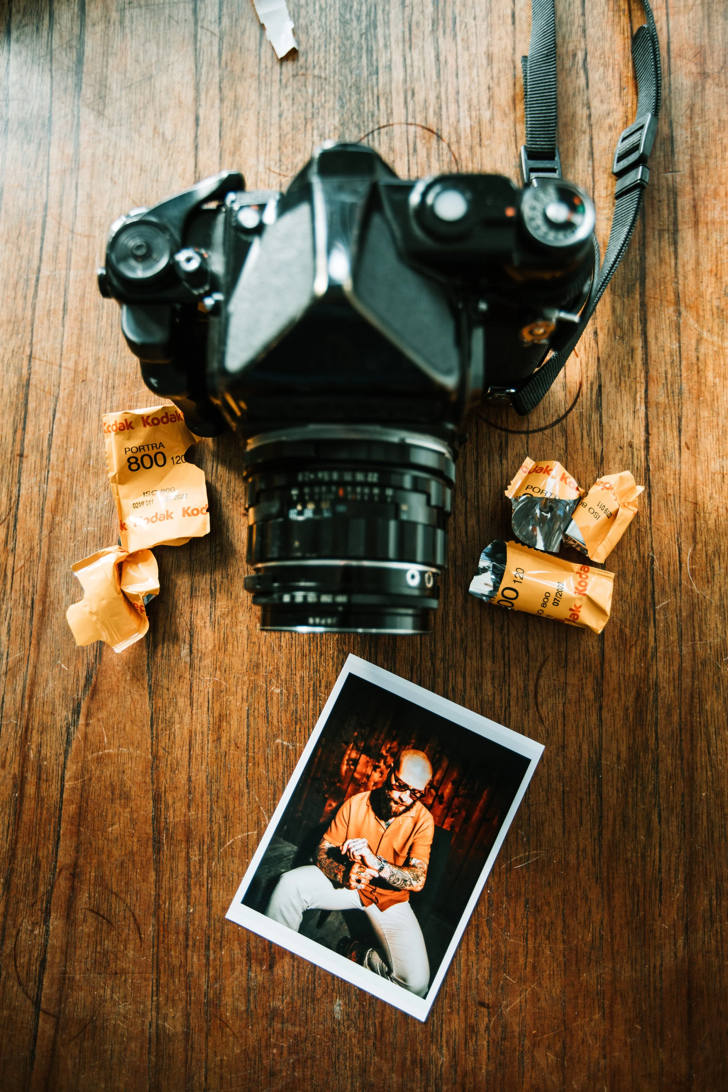 A vintage camera with a telephoto lens on a wooden surface. Surrounding the camera are crumpled Kodak film rolls and a printed photo of a tattooed man wearing glasses and an orange shirt.