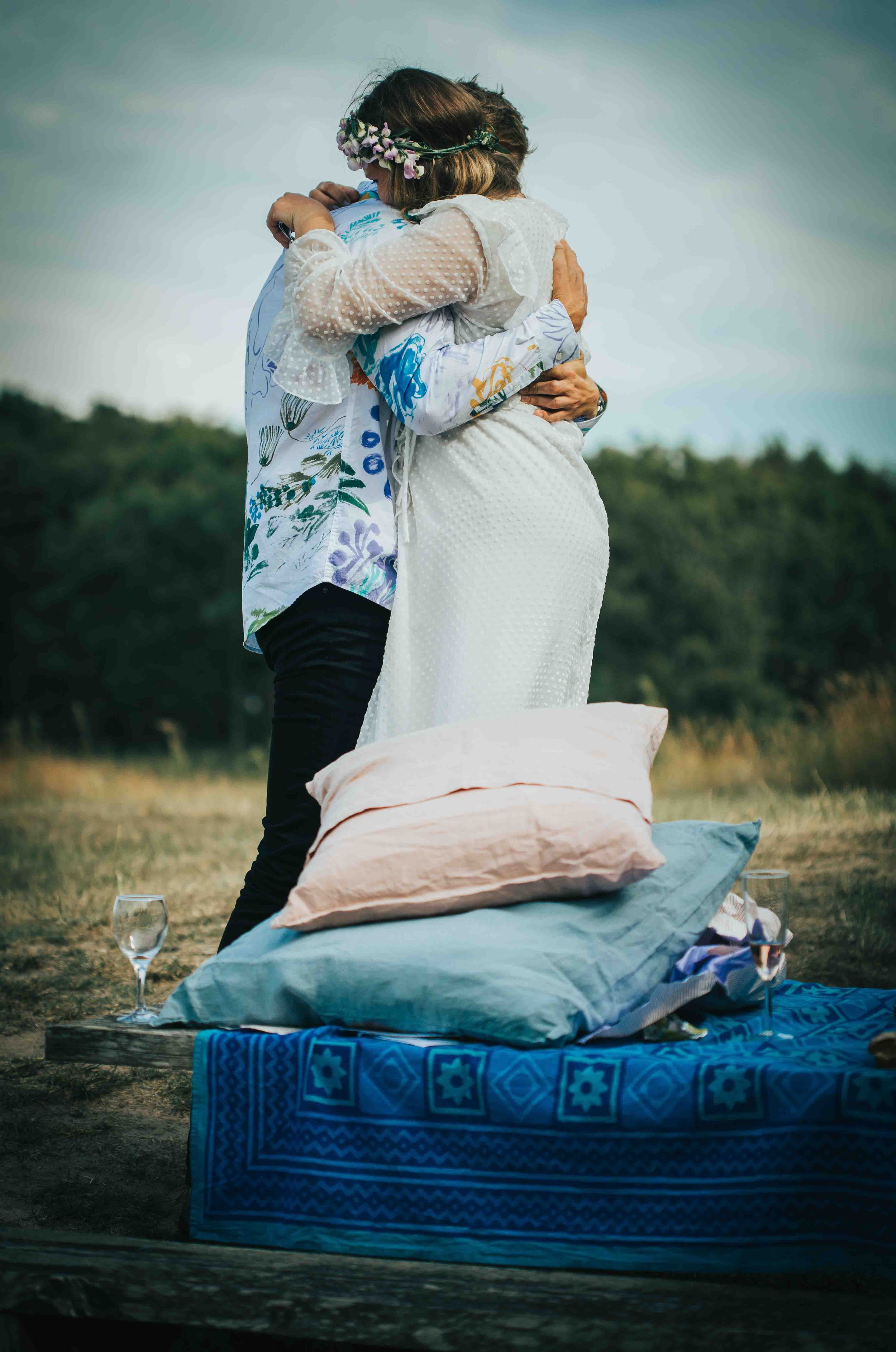 A couple hugging during a romantic outdoor picnic or dance, with pillows and glasses of wine, on a blanket in a natural setting.