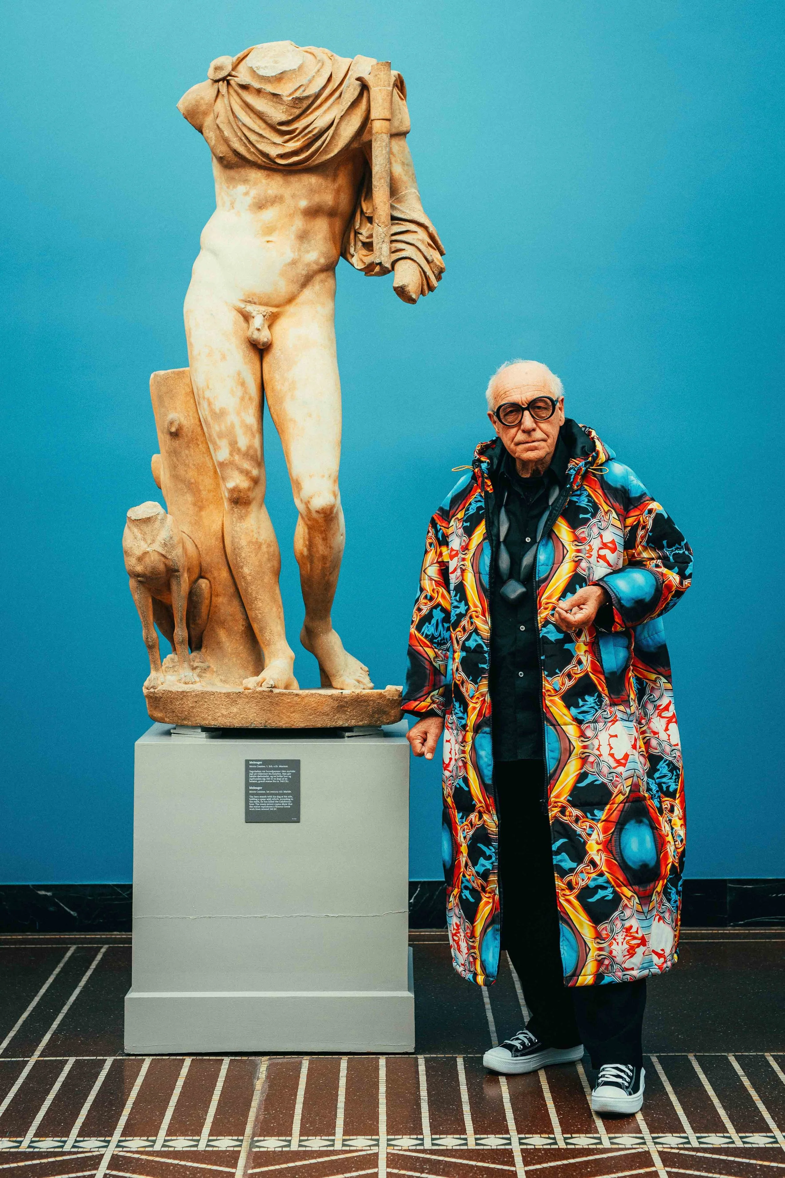 An elderly man wearing a colorful, patterned jacket and black pants standing next to a broken classical sculpture in a museum.
