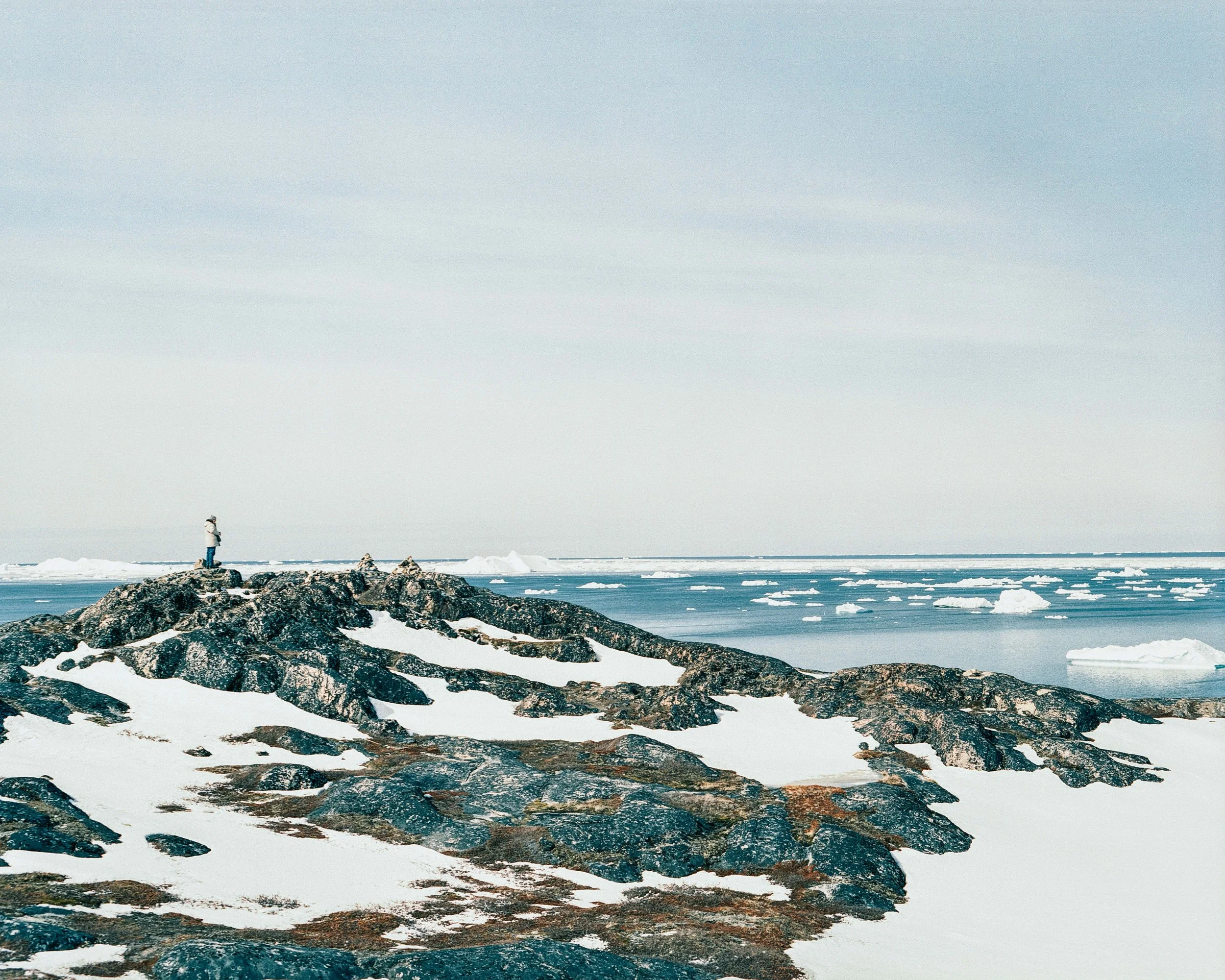 A person standing on a rocky snowy shore looking at the icy ocean with floating icebergs.