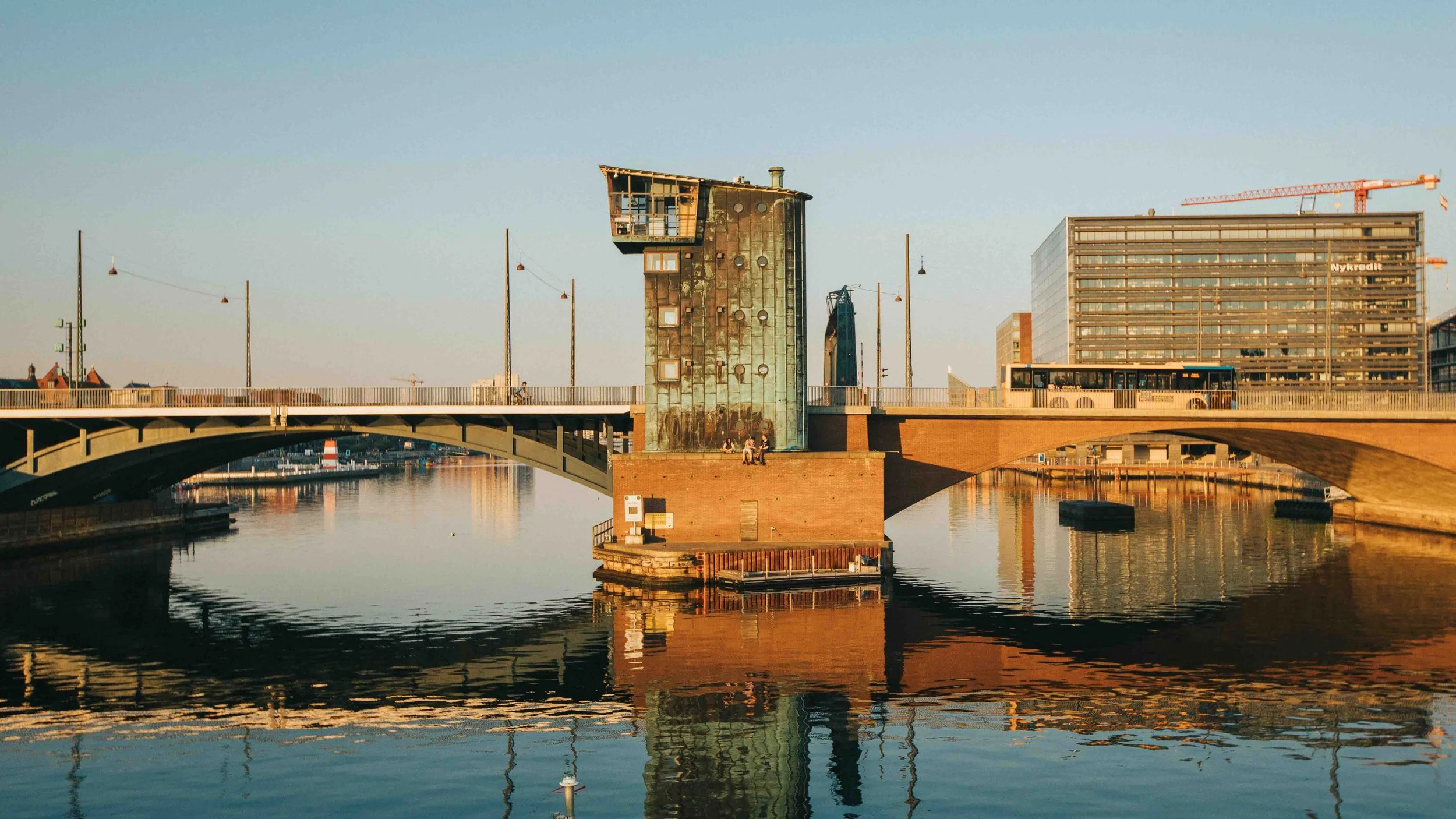 A bridge over a calm river with an unusual modern building and tall buildings in the background, illuminated by warm sunlight.