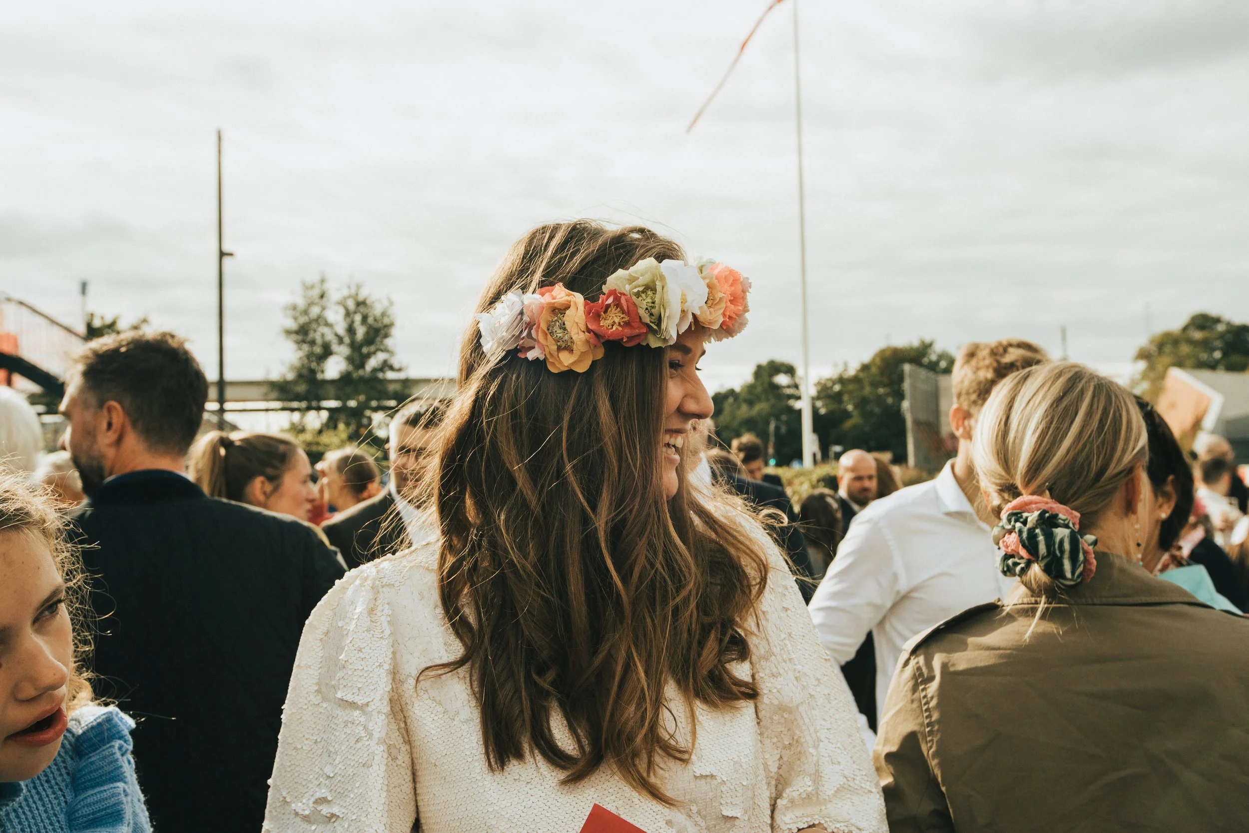 A woman with long brown hair wearing a floral crown and white textured dress, smiling at an outdoor gathering surrounded by people.
