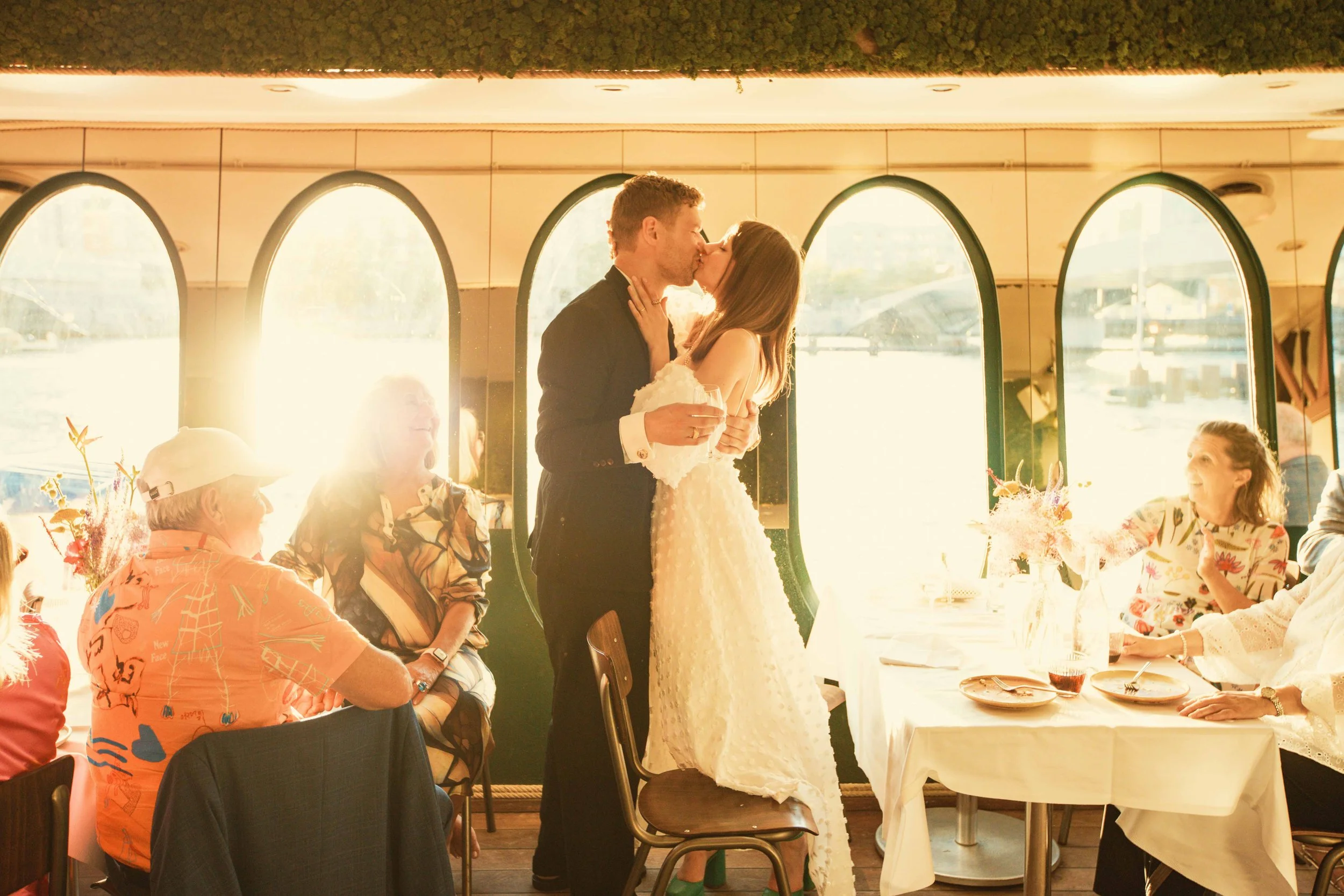 A couple kissing during their wedding reception, surrounded by seated guests at a table with floral decorations, in a bright, sunlit venue with large arched windows.