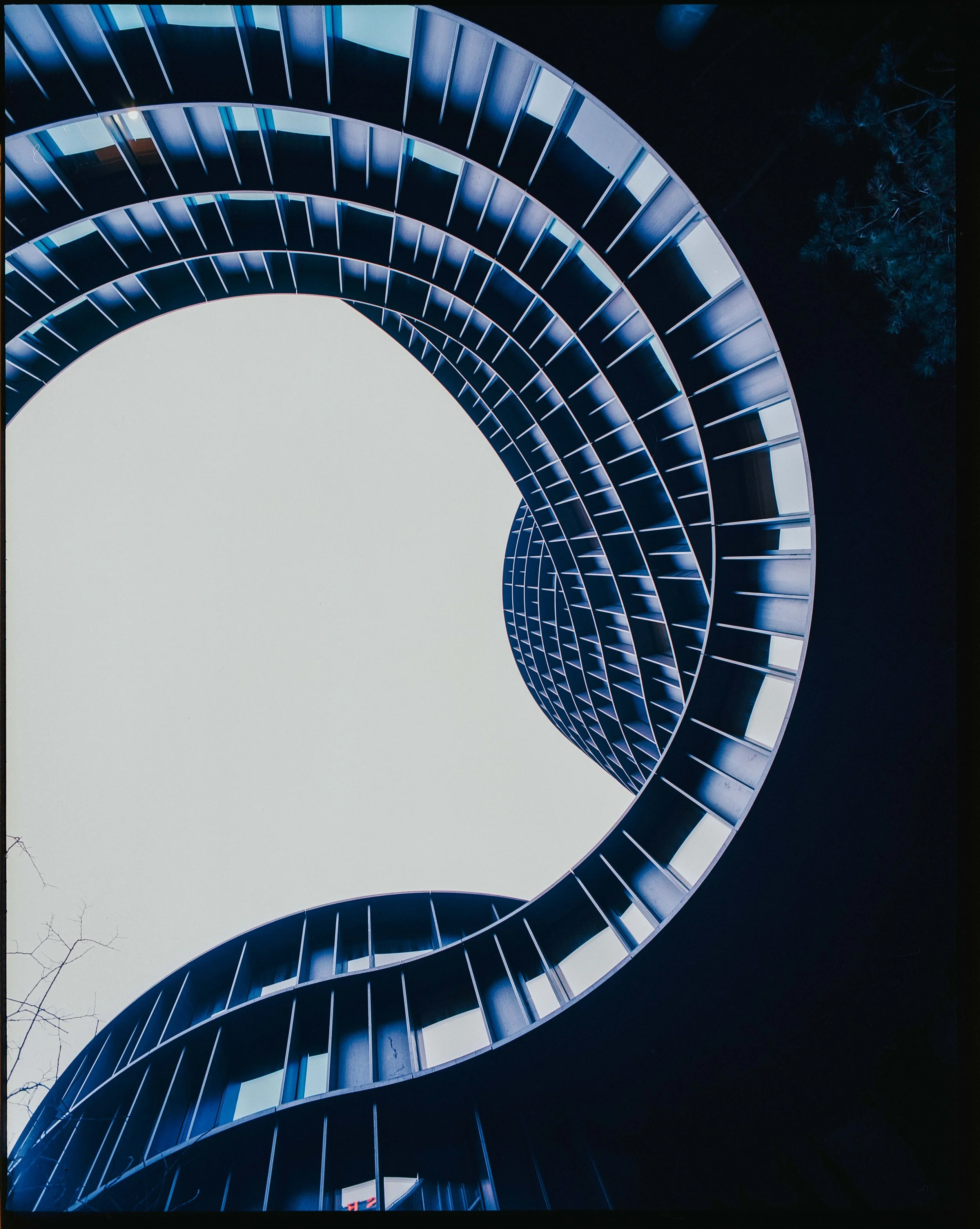 Looking up at a modern spiral staircase with dark blue railings and open sides, set against a bright sky and some trees in the background.