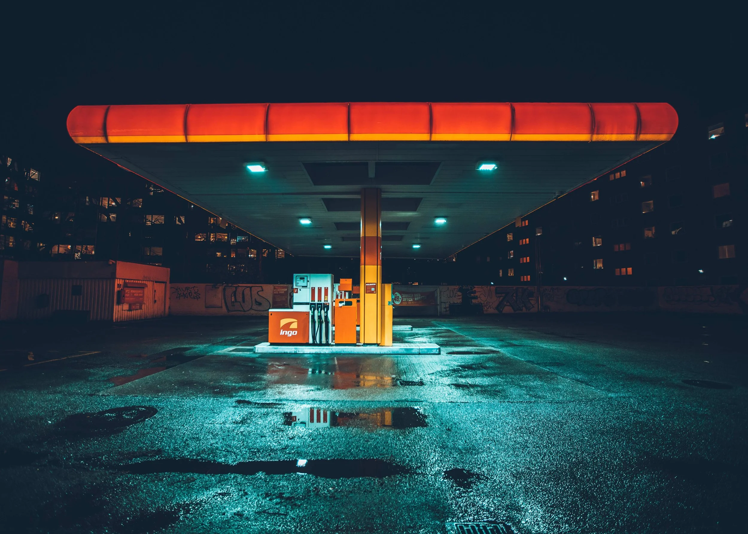 Empty urban gas station at night with wet pavement, illuminated by overhead lights, graffiti on the surrounding walls, and a dark sky.