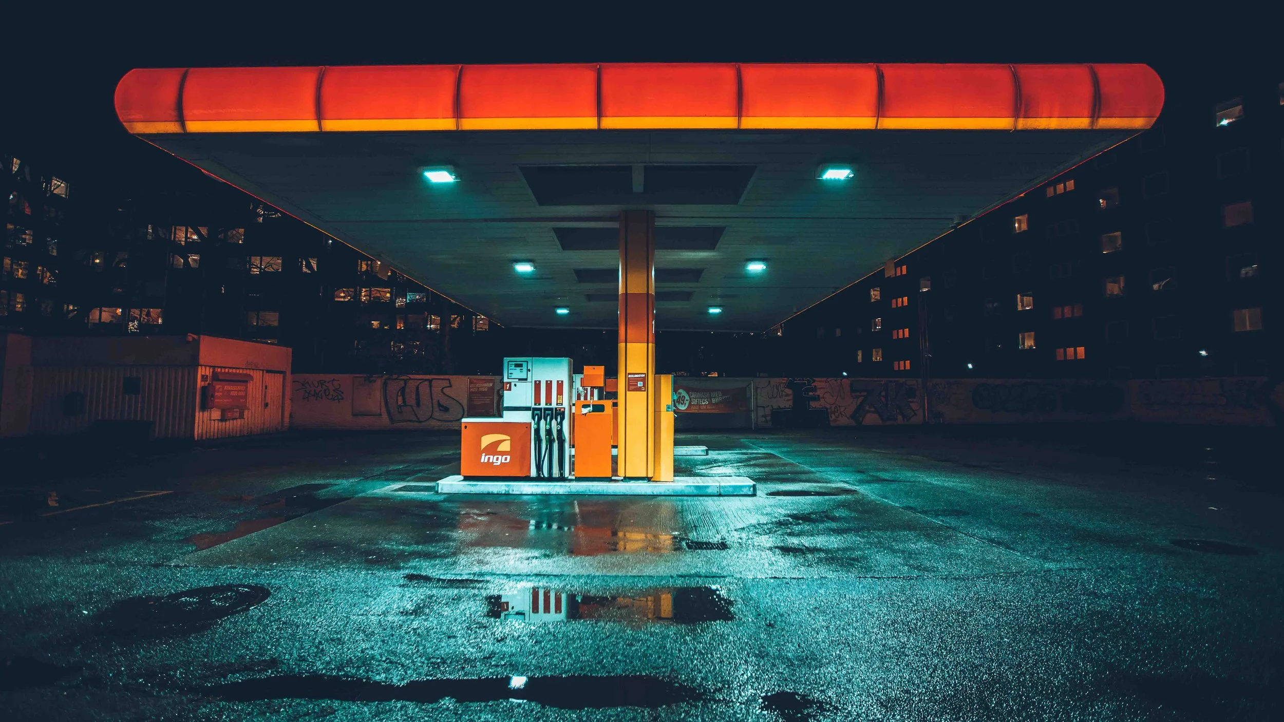 Empty gas station at night with wet pavement reflecting lights, graffiti on the walls, and no cars or people present.