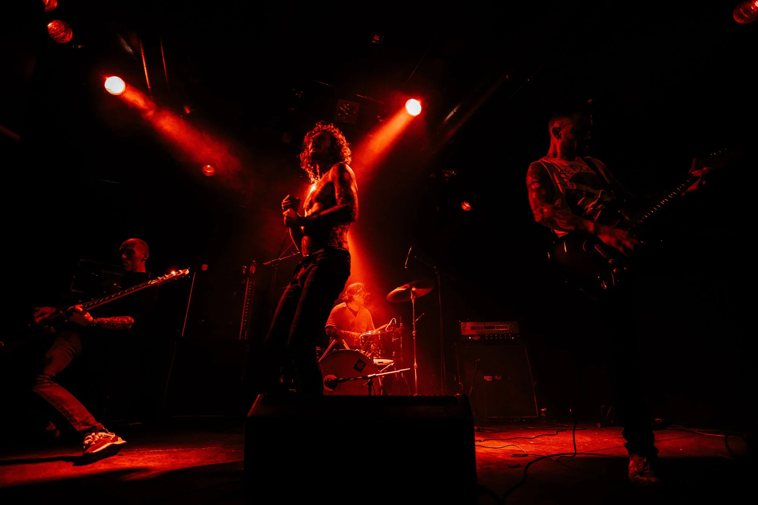 Band performing on stage with red lighting, featuring a female singer, a guitarist, a bassist, and a drummer.