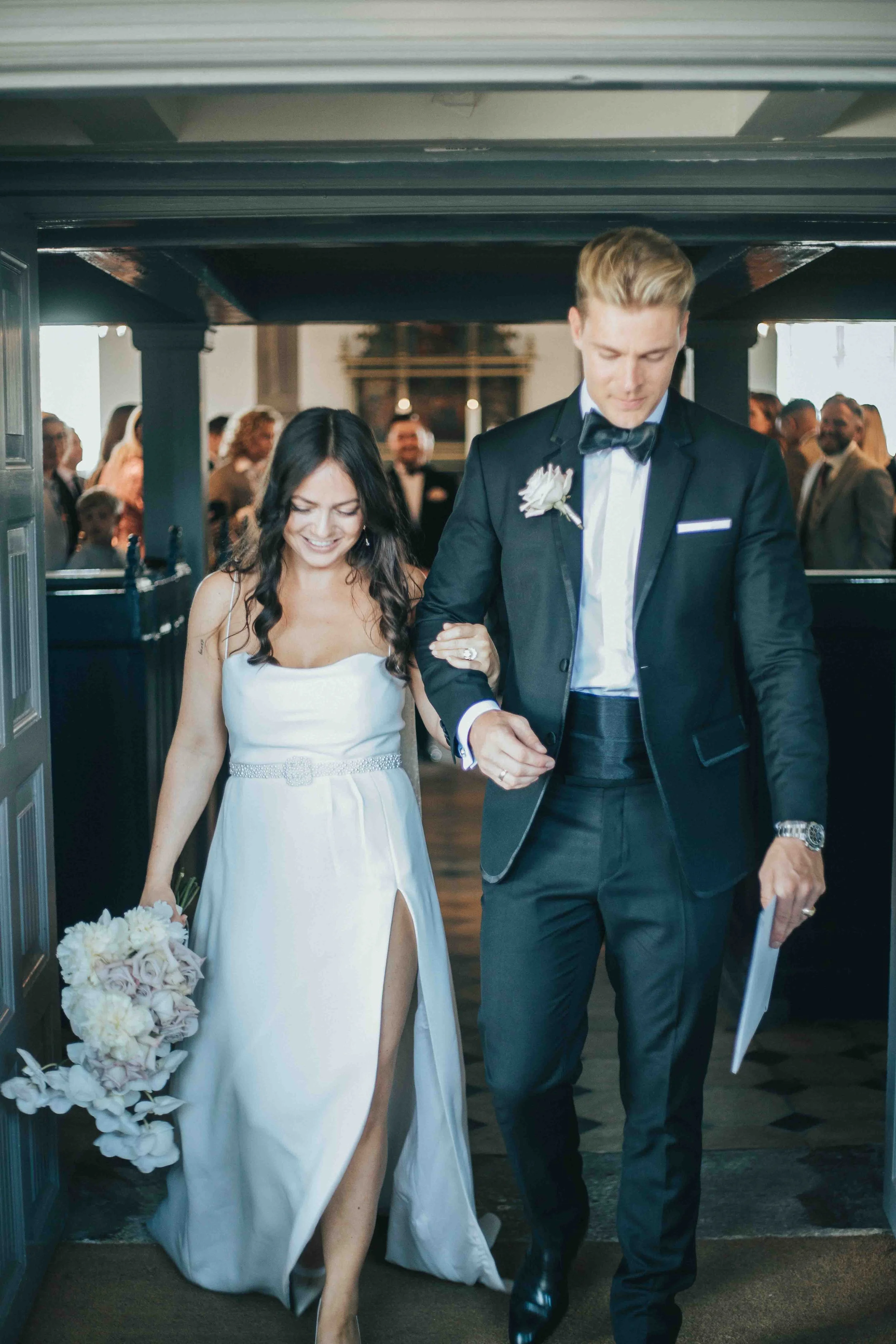 A bride and groom walking together into a wedding venue, surrounded by guests, with the bride holding a bouquet and wearing a white dress, and the groom in a black tuxedo.