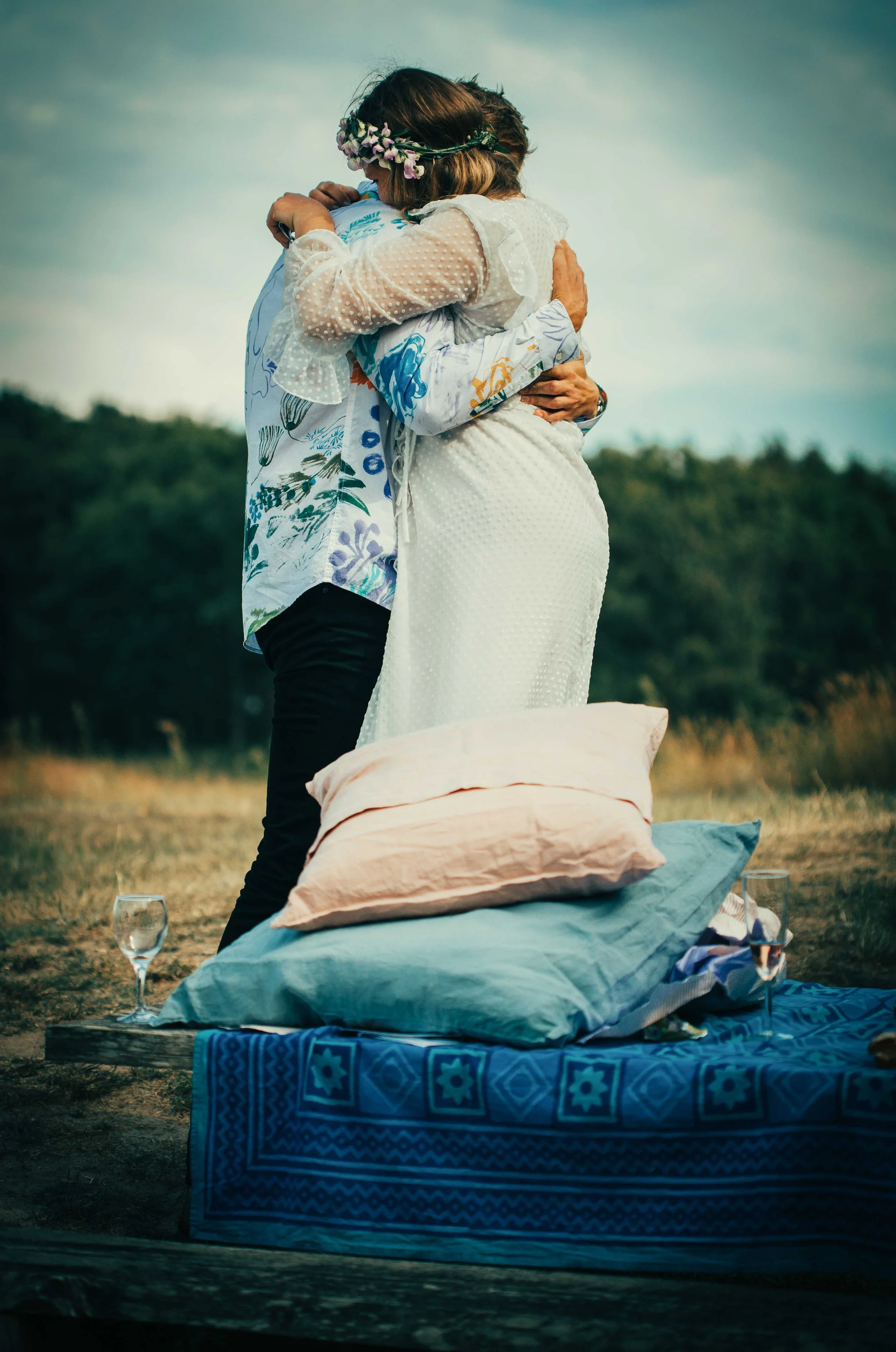 A couple shares a kiss in an outdoor setting with pillows and drinks on a blanket, celebrating a romantic moment.