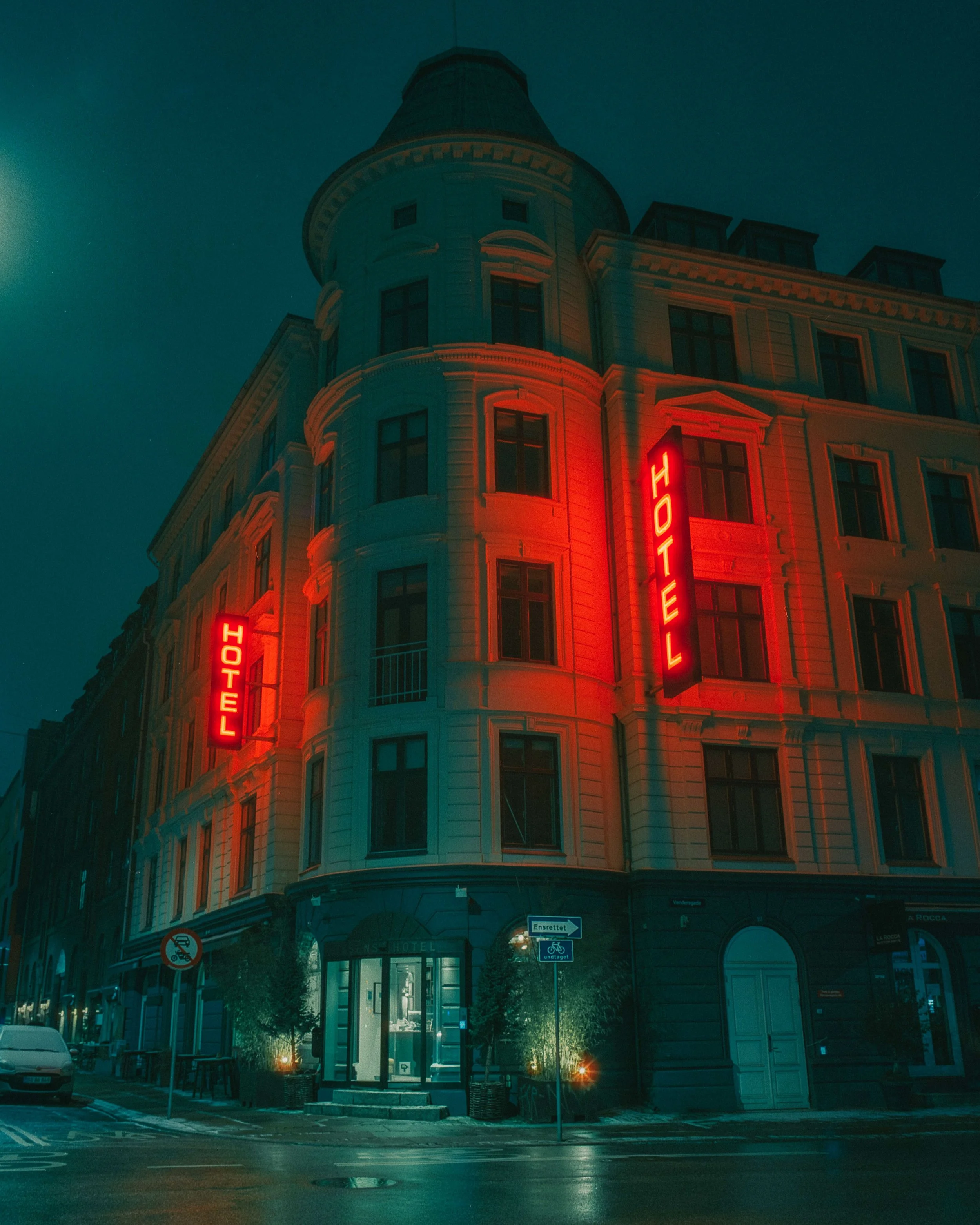 A multi-story hotel building at night with a distinctive European architecture style, illuminated by two vertical red neon signs reading 'HOTEL' on either side of the building. The entrance is lit and surrounded by small trees and street signs.
