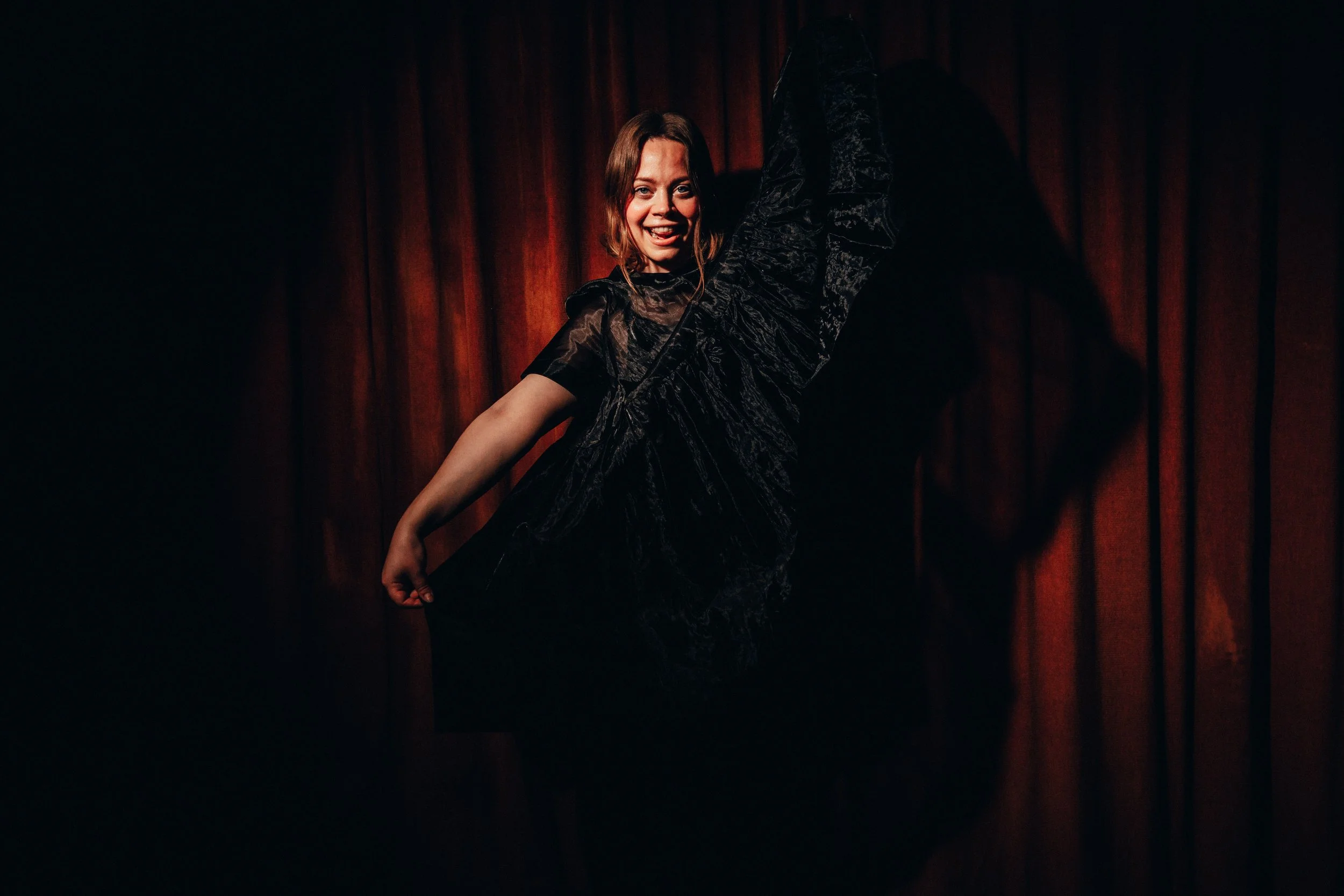 A woman with brown hair in a black dress standing in front of red curtains, smiling and holding one arm high with a shadow cast on the curtain.