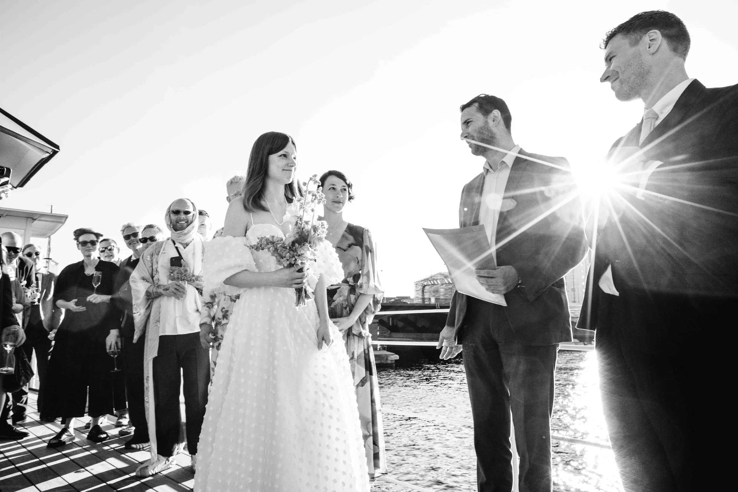 A black and white photo of a wedding ceremony on a boat, with a bride holding a bouquet facing the groom and officiant, surrounded by guests, with the sun setting over the water in the background.