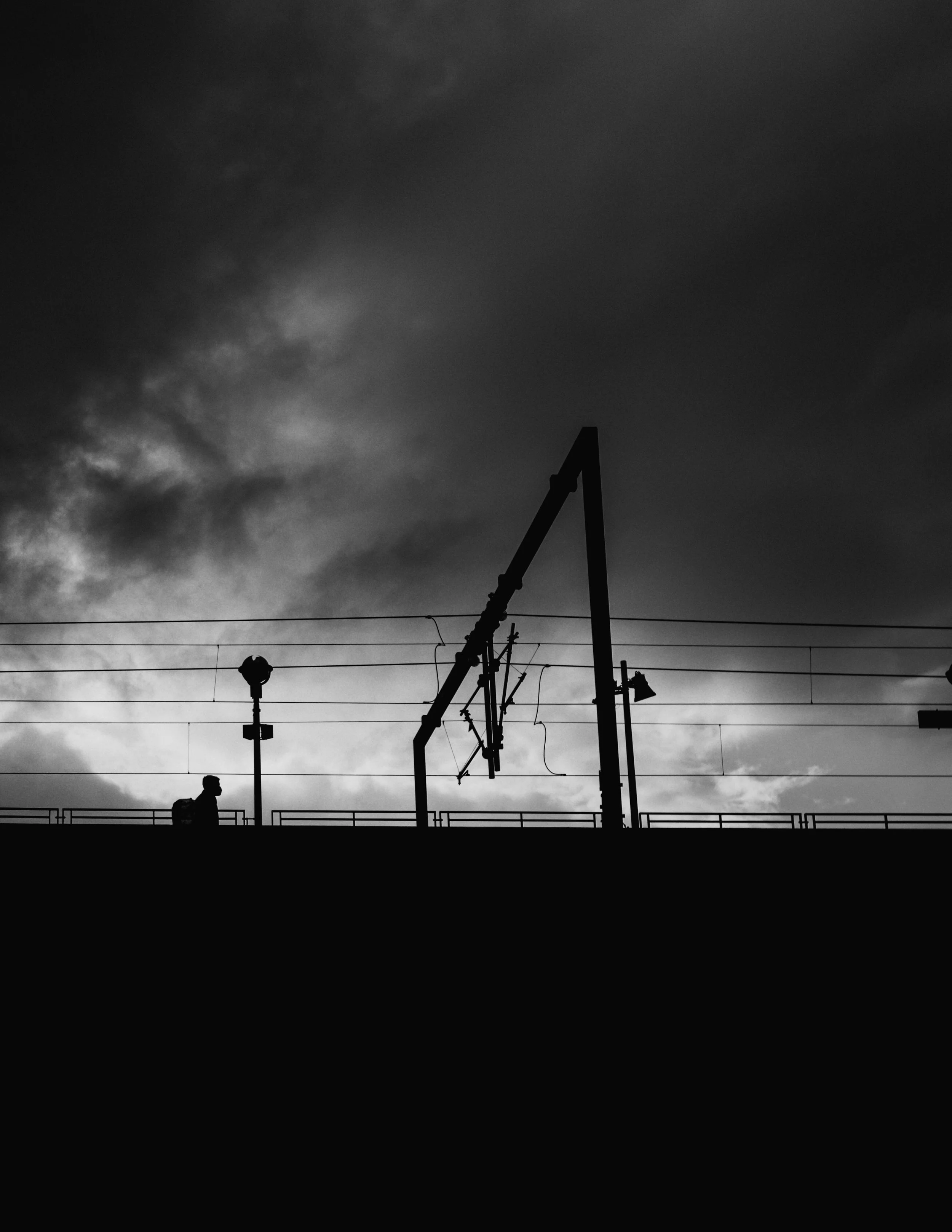 Silhouette of a person with a backpack walking on a train station platform, with railway tracks, overhead electrical lines, and a cloudy sky in the background.