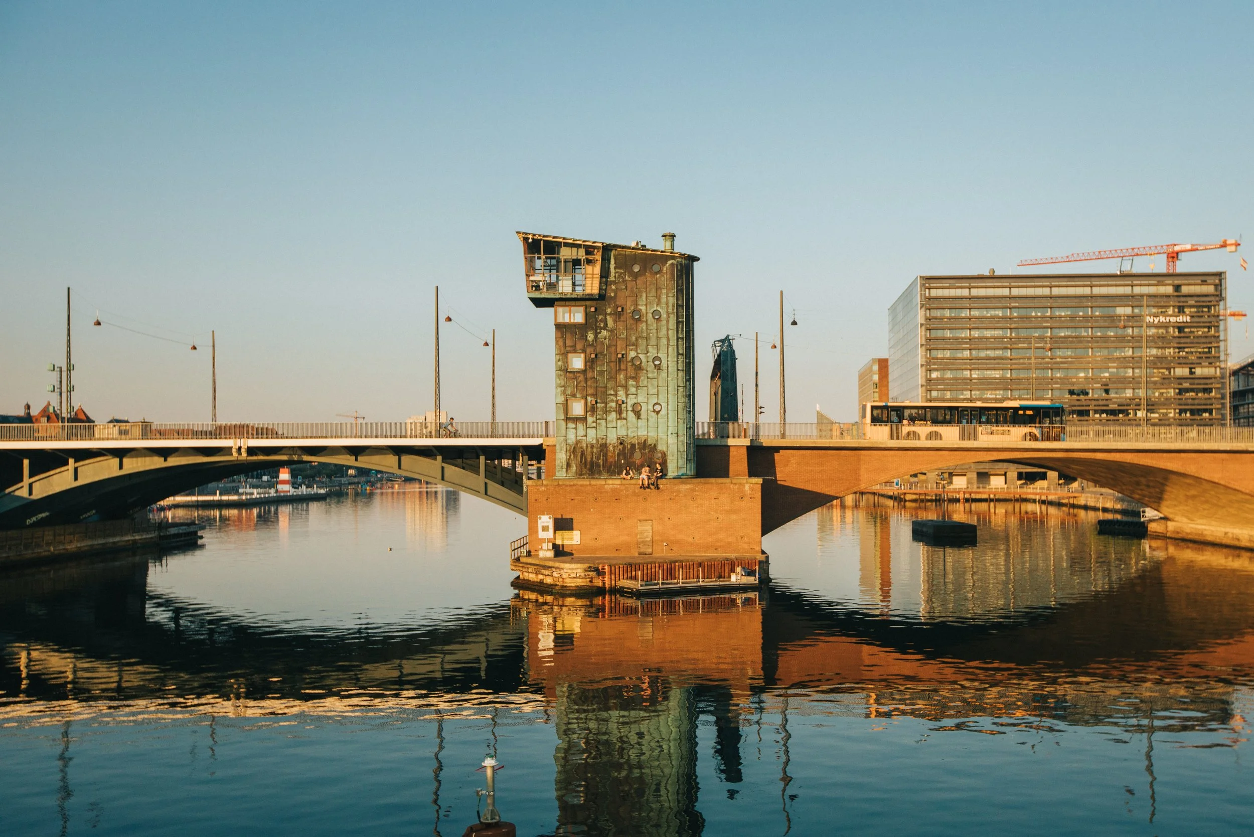 A bridge over a calm river with a modern, irregularly shaped building on a brick base in the center. The building has a distinctive angular top and weathered greenish exterior. Reflections of the structures are visible in the water. Modern high-rises