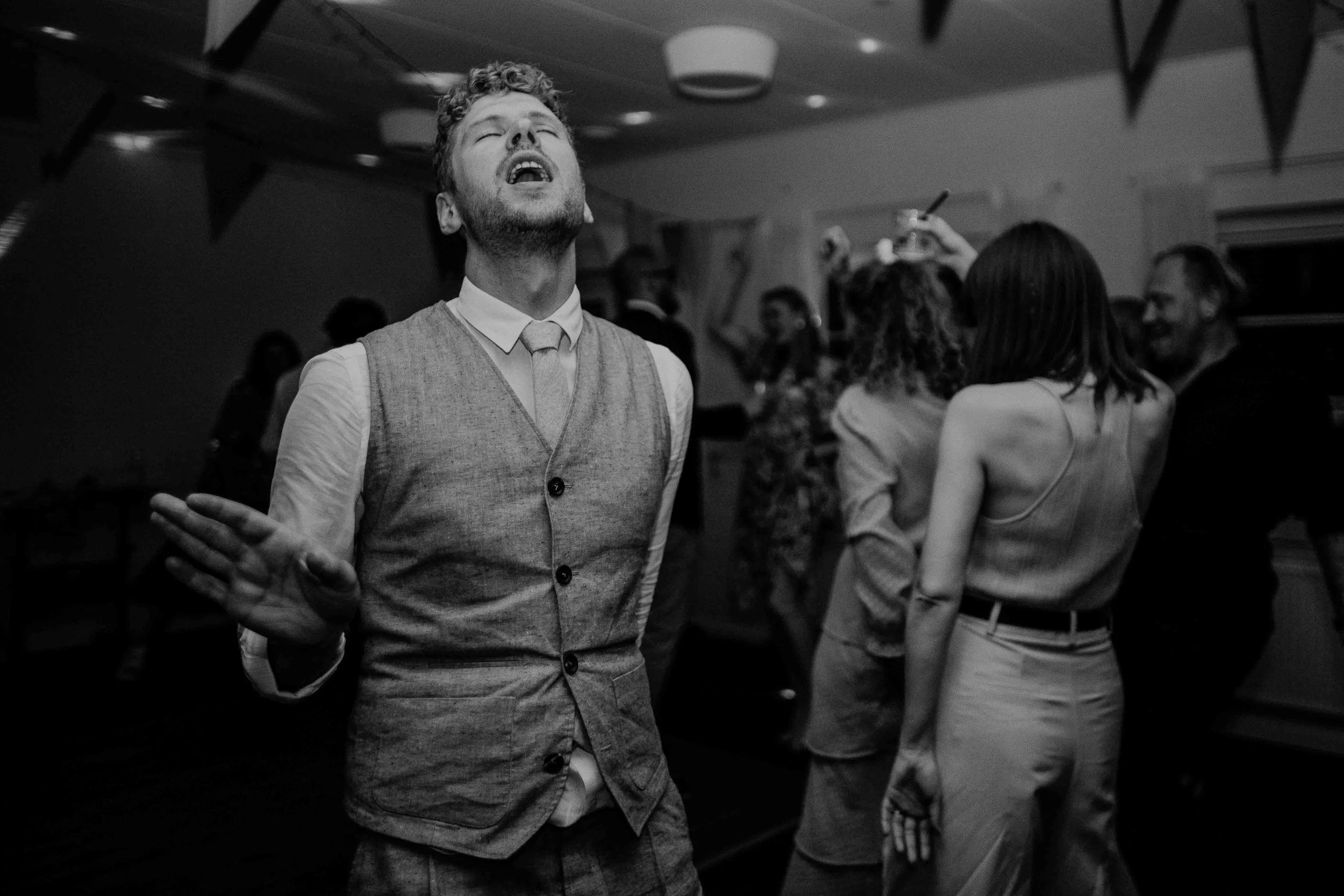 A man in a vest and tie at a party with his eyes closed and hand raised, surrounded by people dancing and socializing in a decorated room.