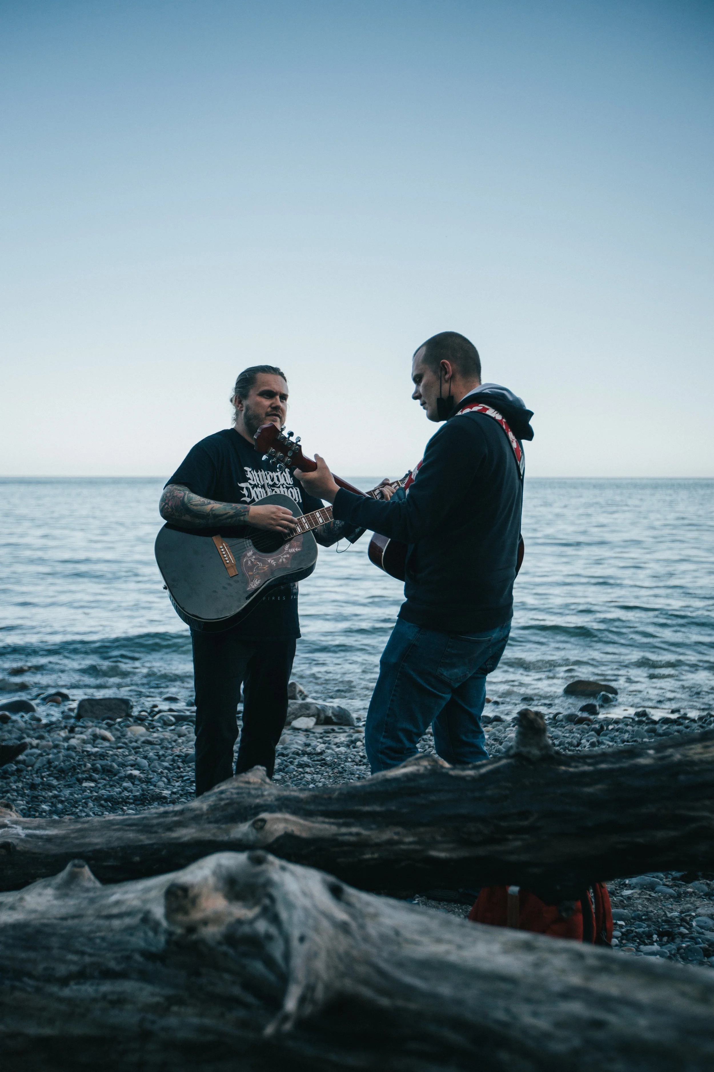 Two men playing acoustic guitars on a rocky beach near the ocean during dusk.