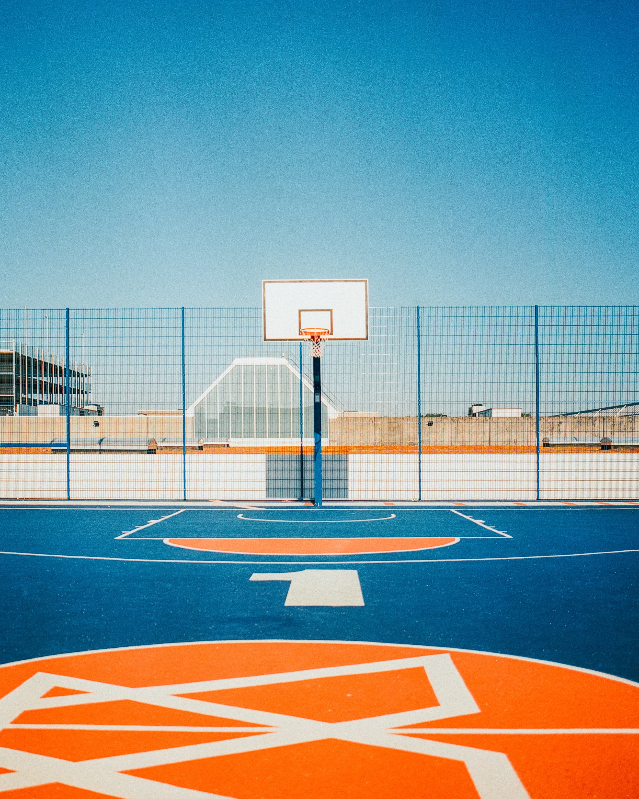 Empty outdoor basketball court with a hoop, surrounded by a blue fence, under a clear blue sky.