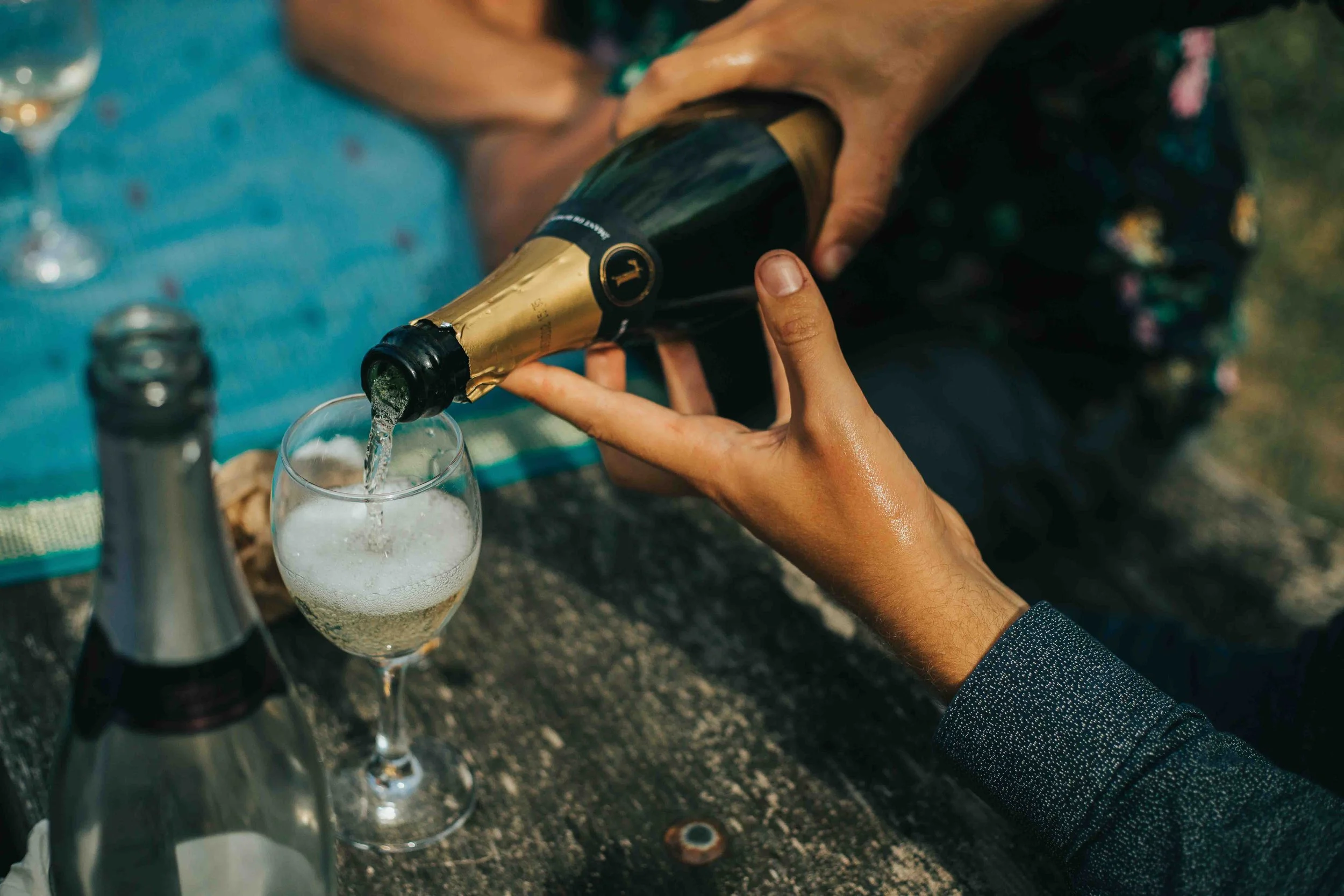 Person pours champagne into a glass at an outdoor gathering, with other bottles and glasses on a wooden table.