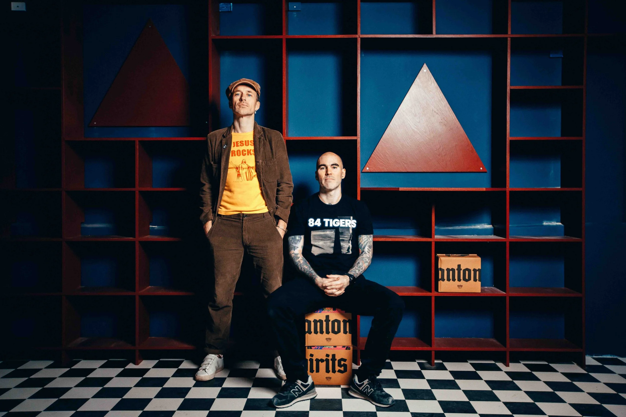 Two men posing in front of a blue wall with red shelves and geometric shapes, one standing and one sitting on a box, in a room with black-and-white checkered flooring.