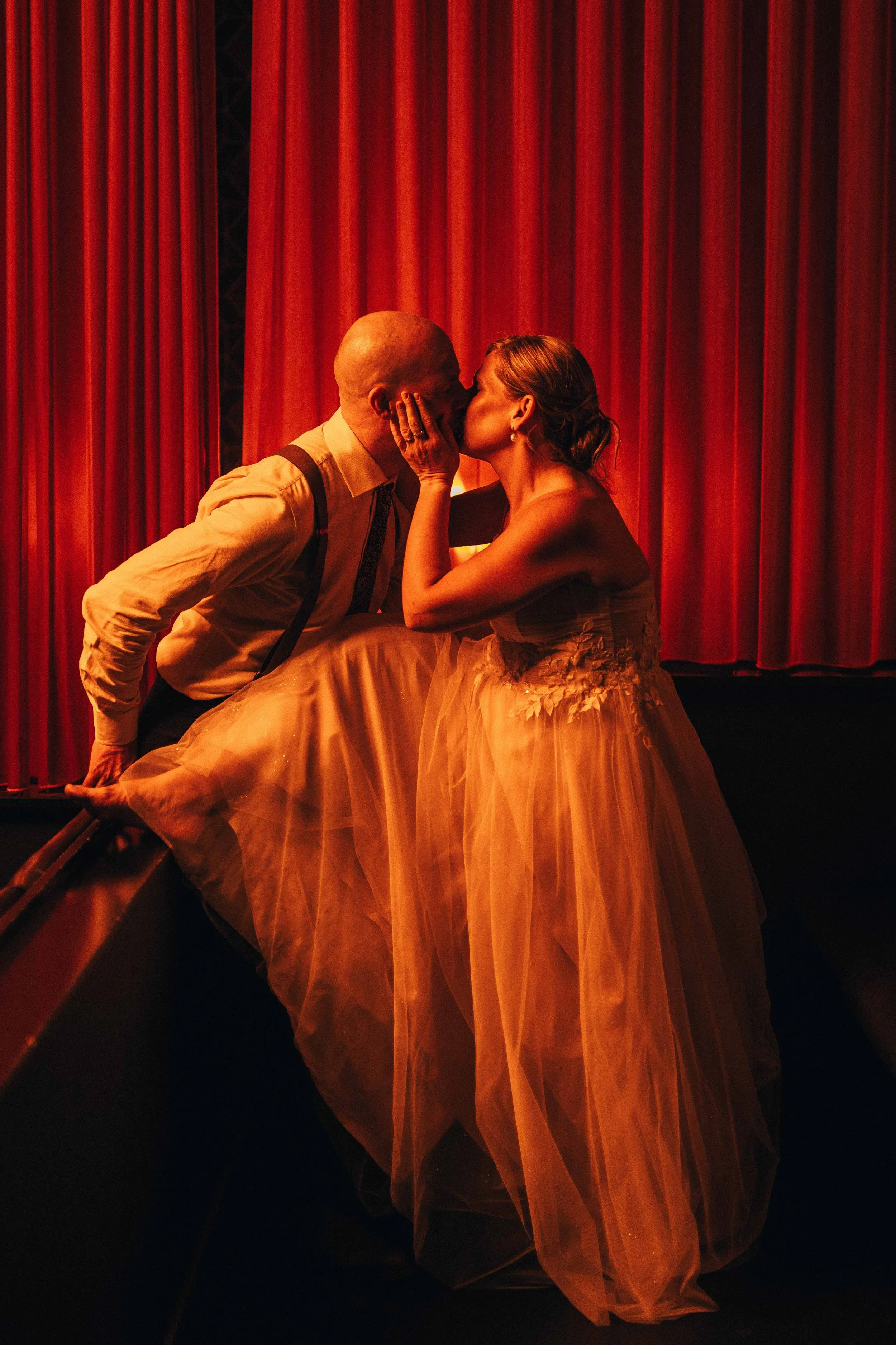A couple shares a kiss backstage at a theater, with red curtains in the background. The woman is in a strapless dress and the man is in a shirt with suspenders.