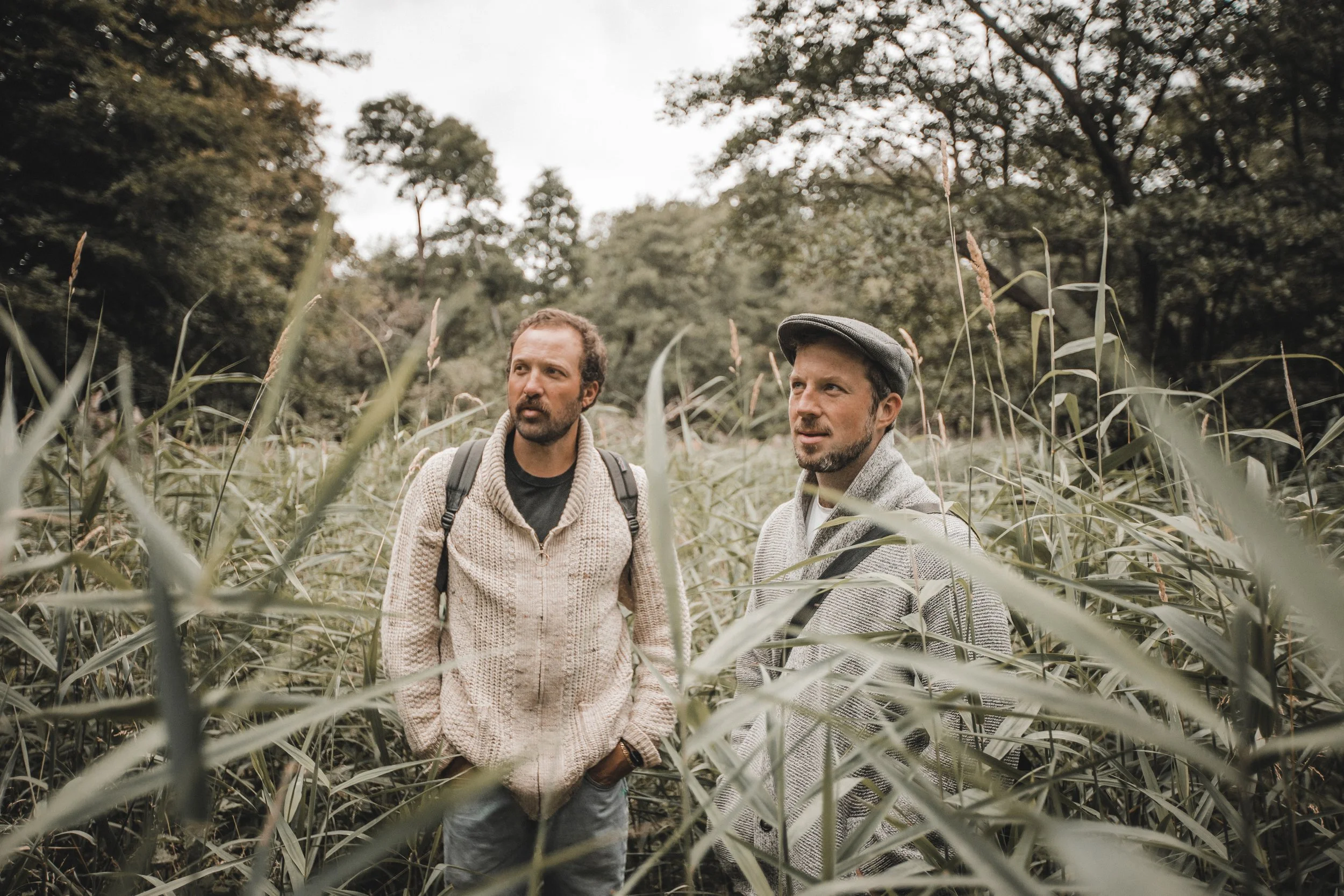 Two men standing outdoors in a field of tall grass with trees in the background, engaged in conversation on an overcast day.