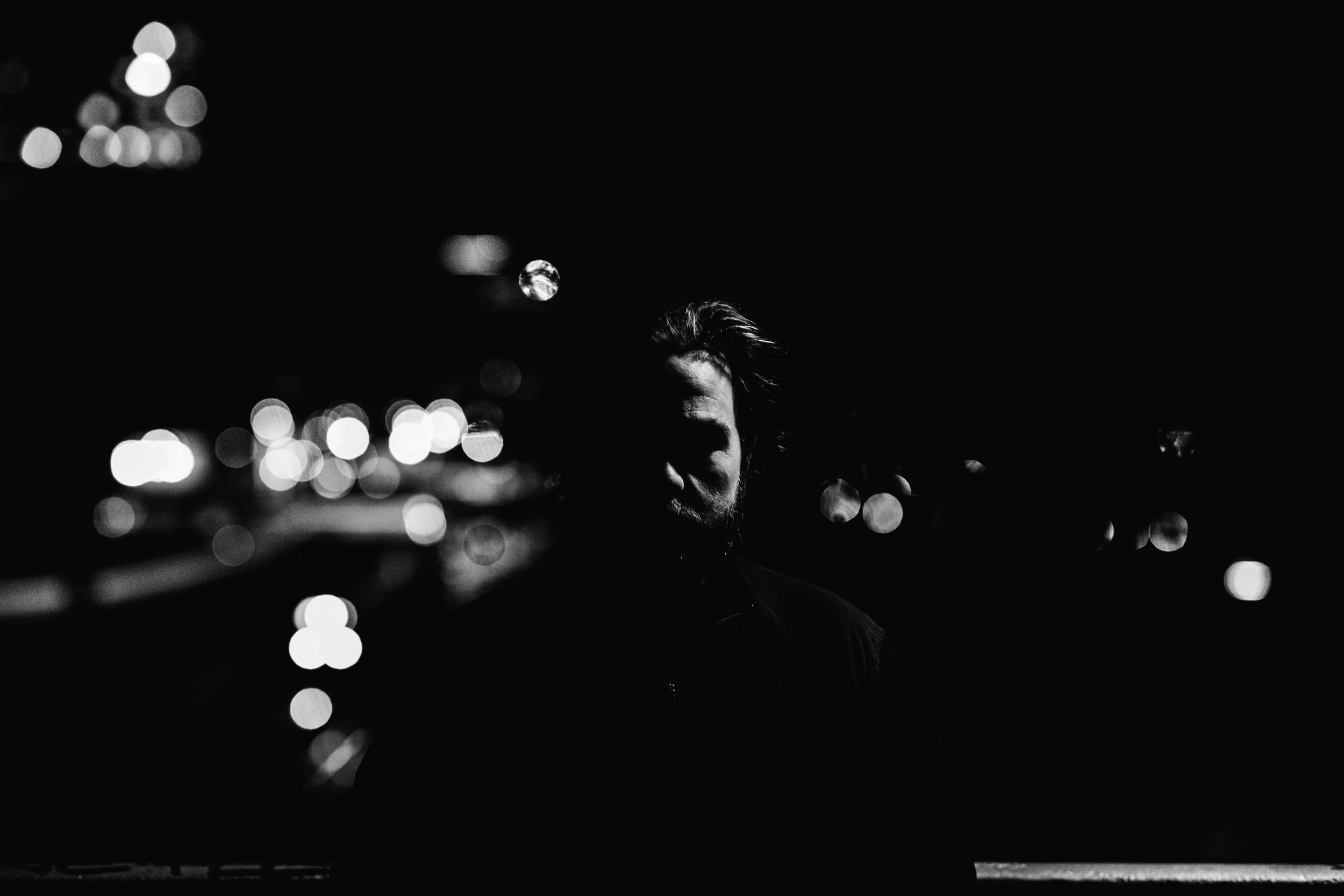 A person with light-colored hair and beard, illuminated from behind, standing outside at night with blurred city lights in the background.