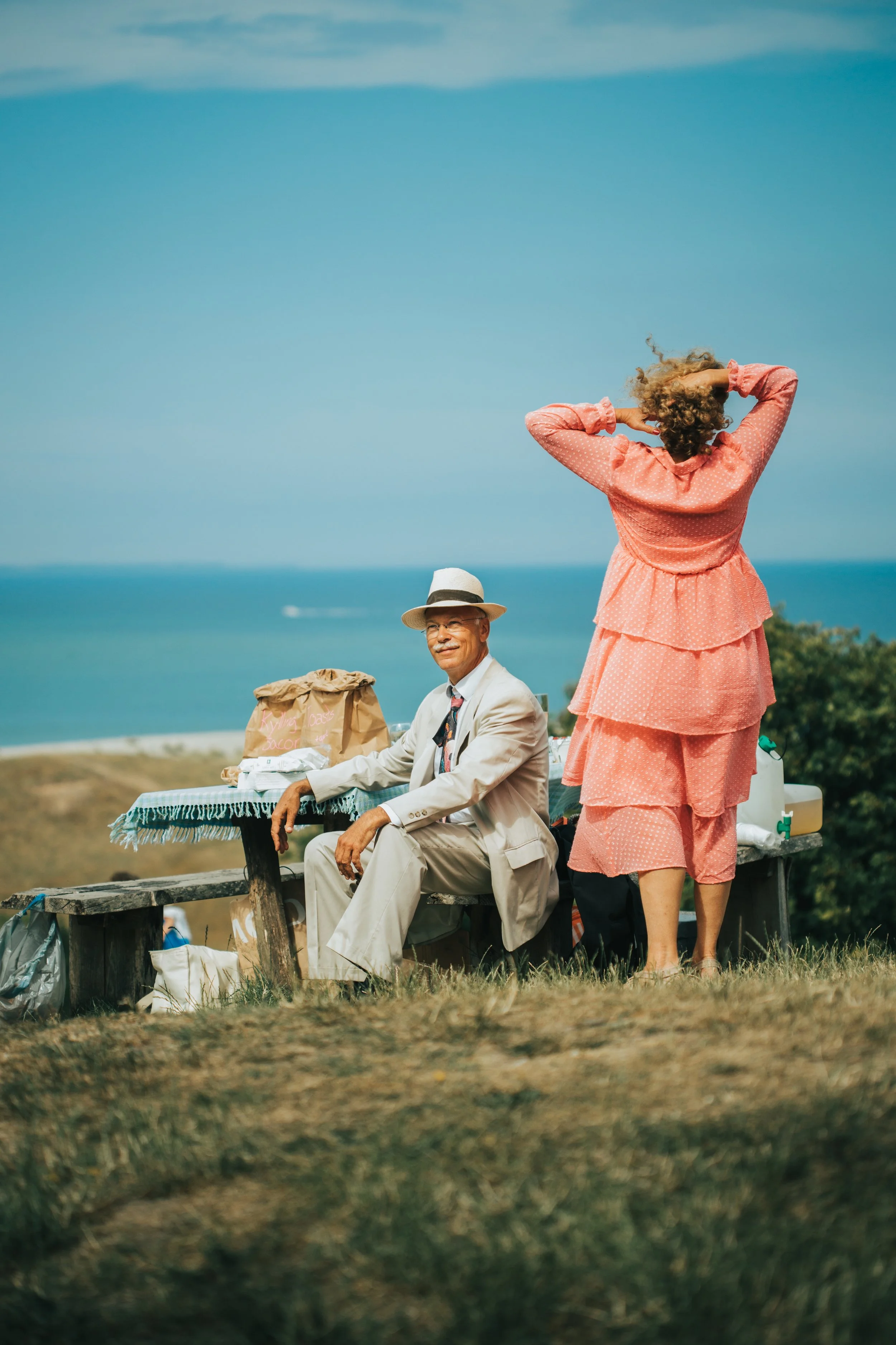 A man in a light-colored suit and straw hat sitting on a picnic bench, with a woman in a pink tiered dress standing behind and adjusting her hair, overlooking a scenic ocean view on a sunny day.