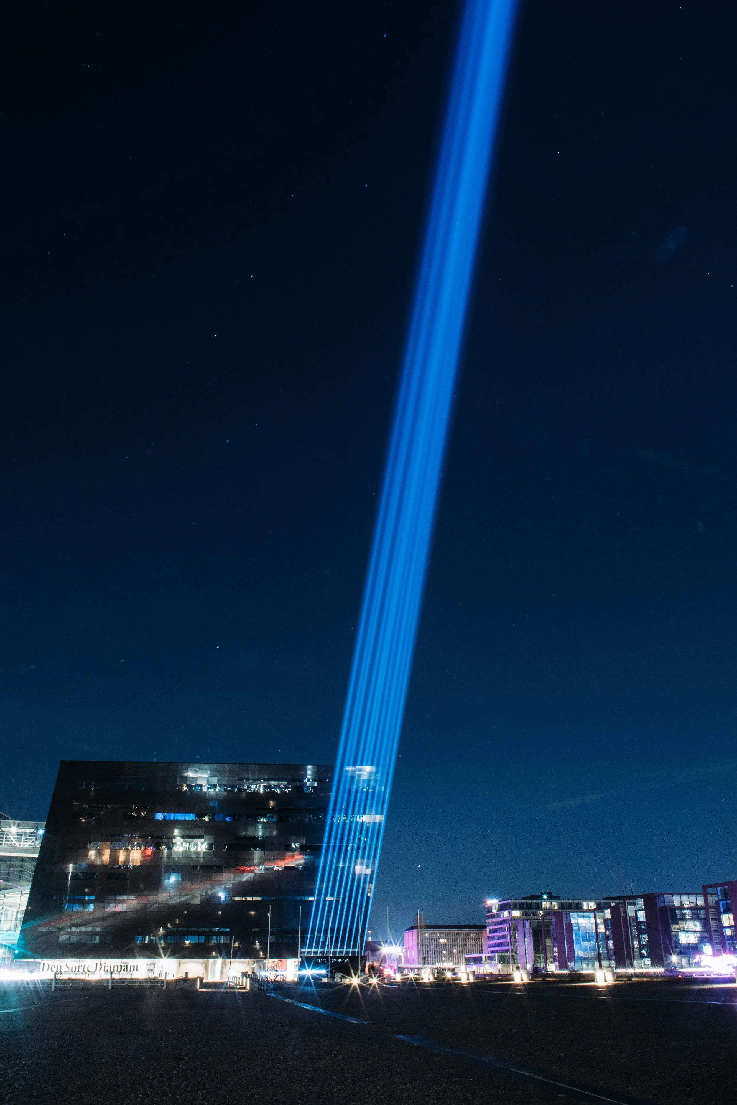 Nighttime cityscape with a beam of blue light projecting into the night sky over modern buildings.