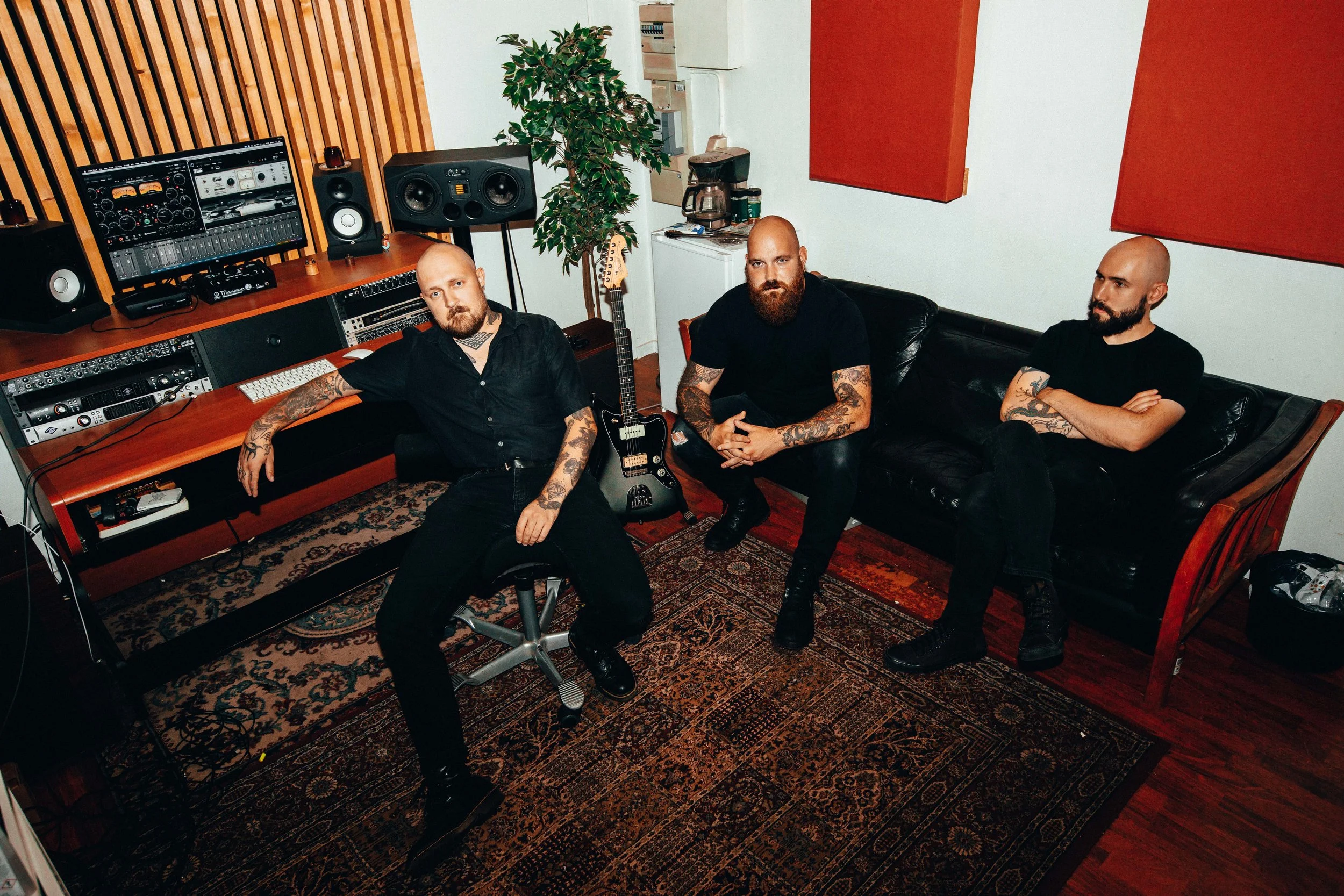 Three men with tattoos sitting in a recording studio, surrounded by audio equipment, a guitar, and red acoustic panels on the wall.
