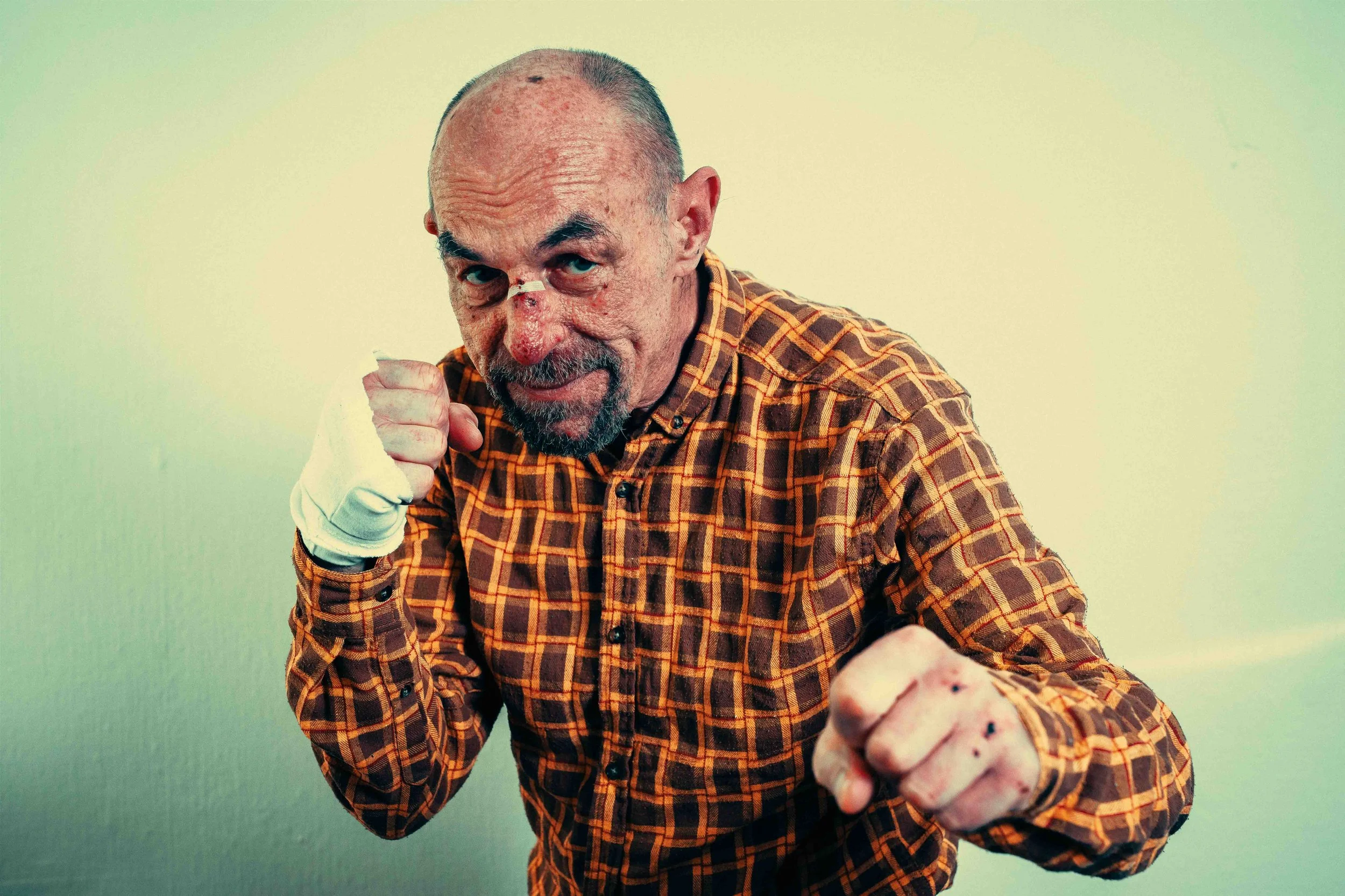 Elderly man with a bandage on his nose, wearing a checkered shirt, posing with fists raised as if ready to fight, standing against a plain background.