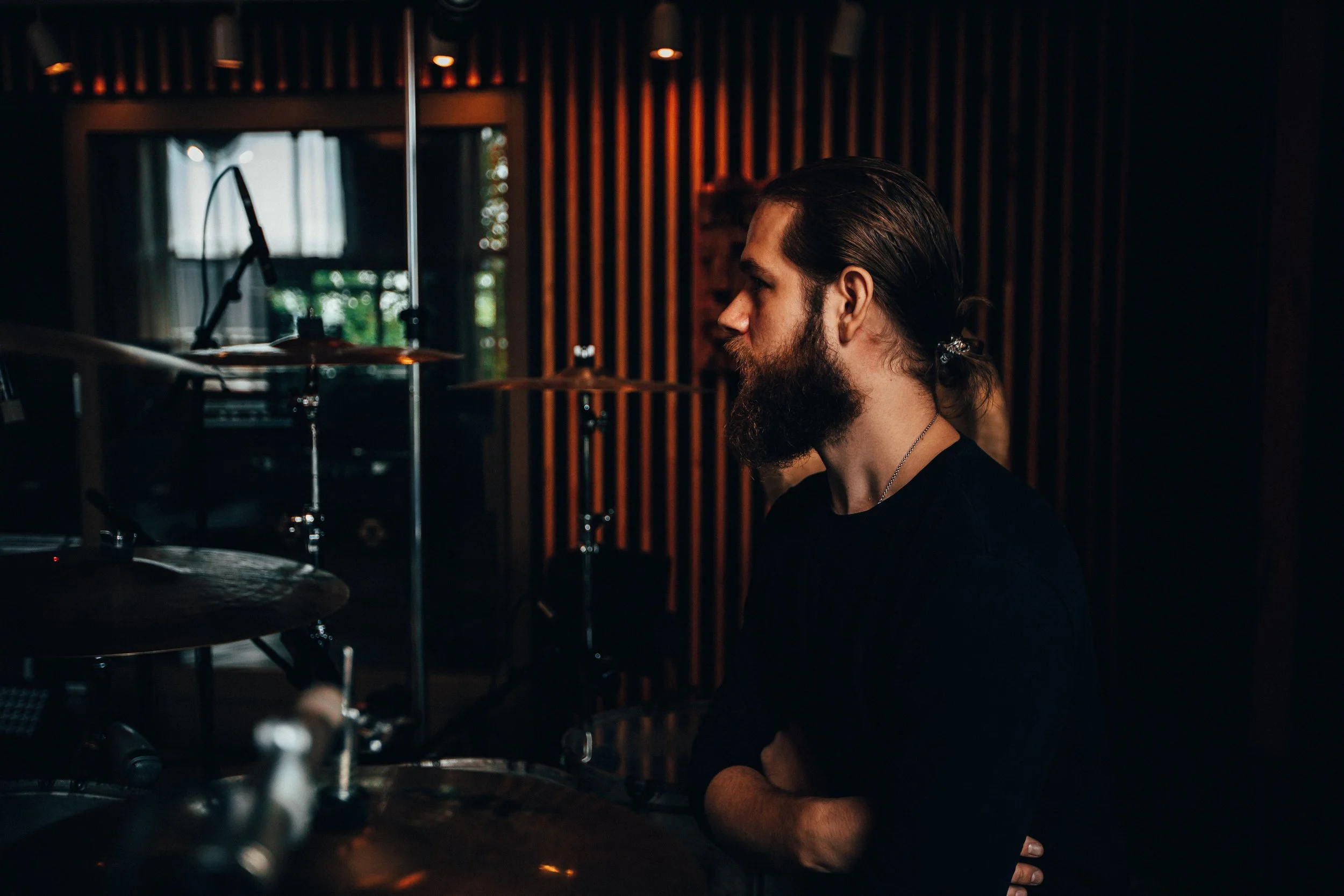 A man with a beard and long hair tied back, sitting in a recording studio, with drums and microphones nearby, dark lighting with wooden wall panels in the background.