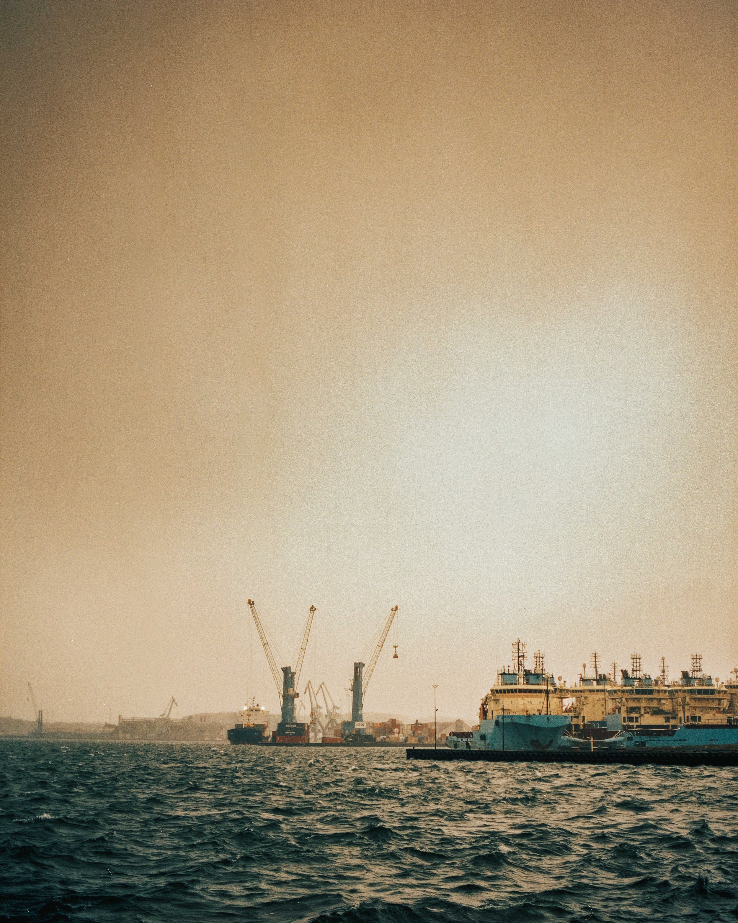 View of a port with ships, cranes, and docked boats during sunset or sunrise.