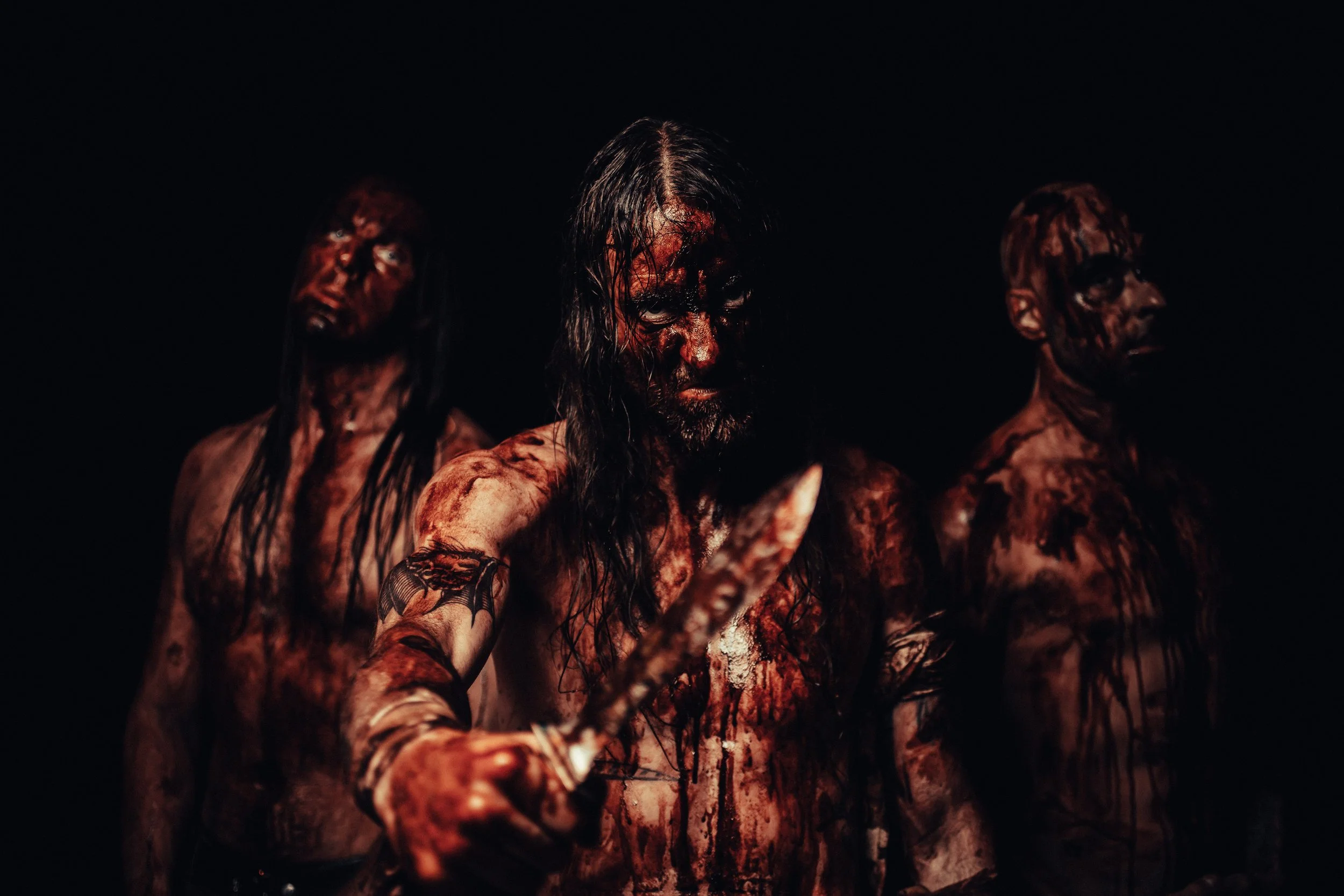 Three people dressed as horror characters with blood on their faces and bodies, one holding a knife, against a dark background.