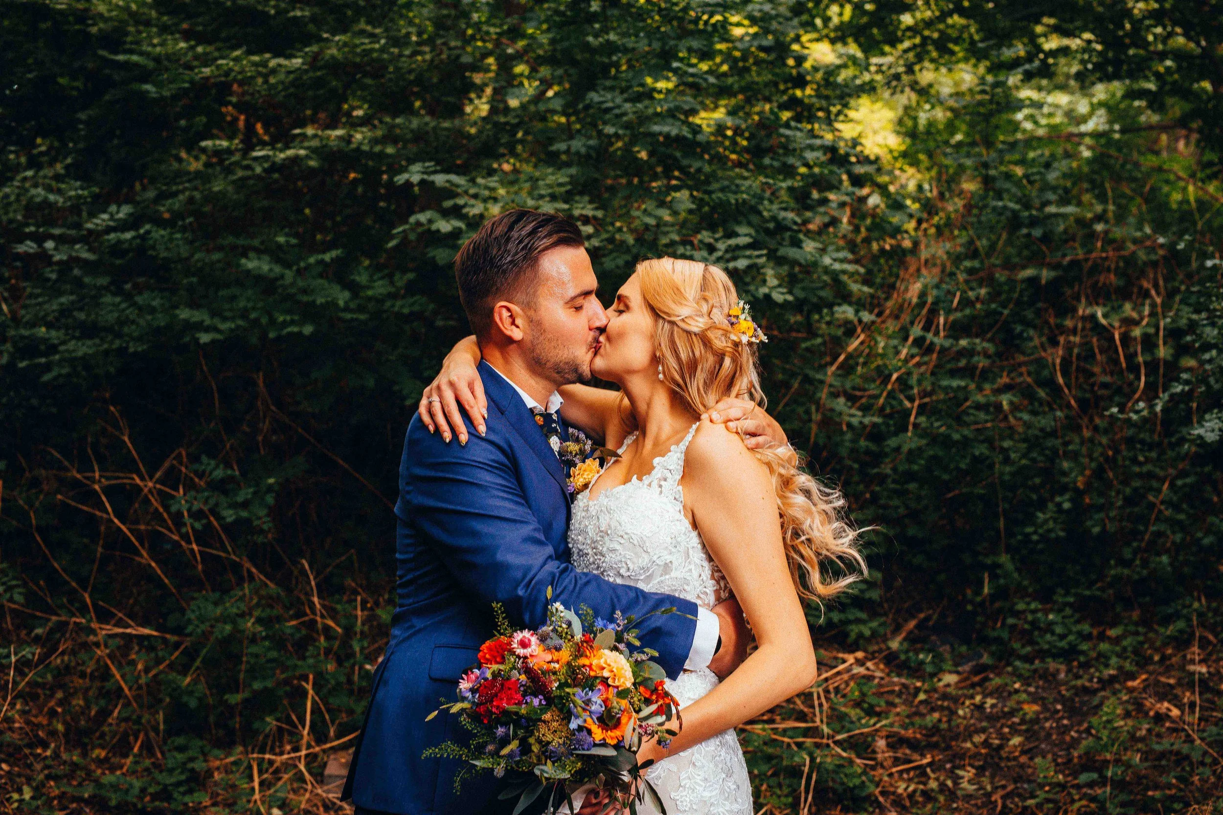 A bride and groom sharing a kiss outdoors in a wooded area, the bride holding a colorful bouquet and wearing a white lace wedding dress, the groom in a blue suit.