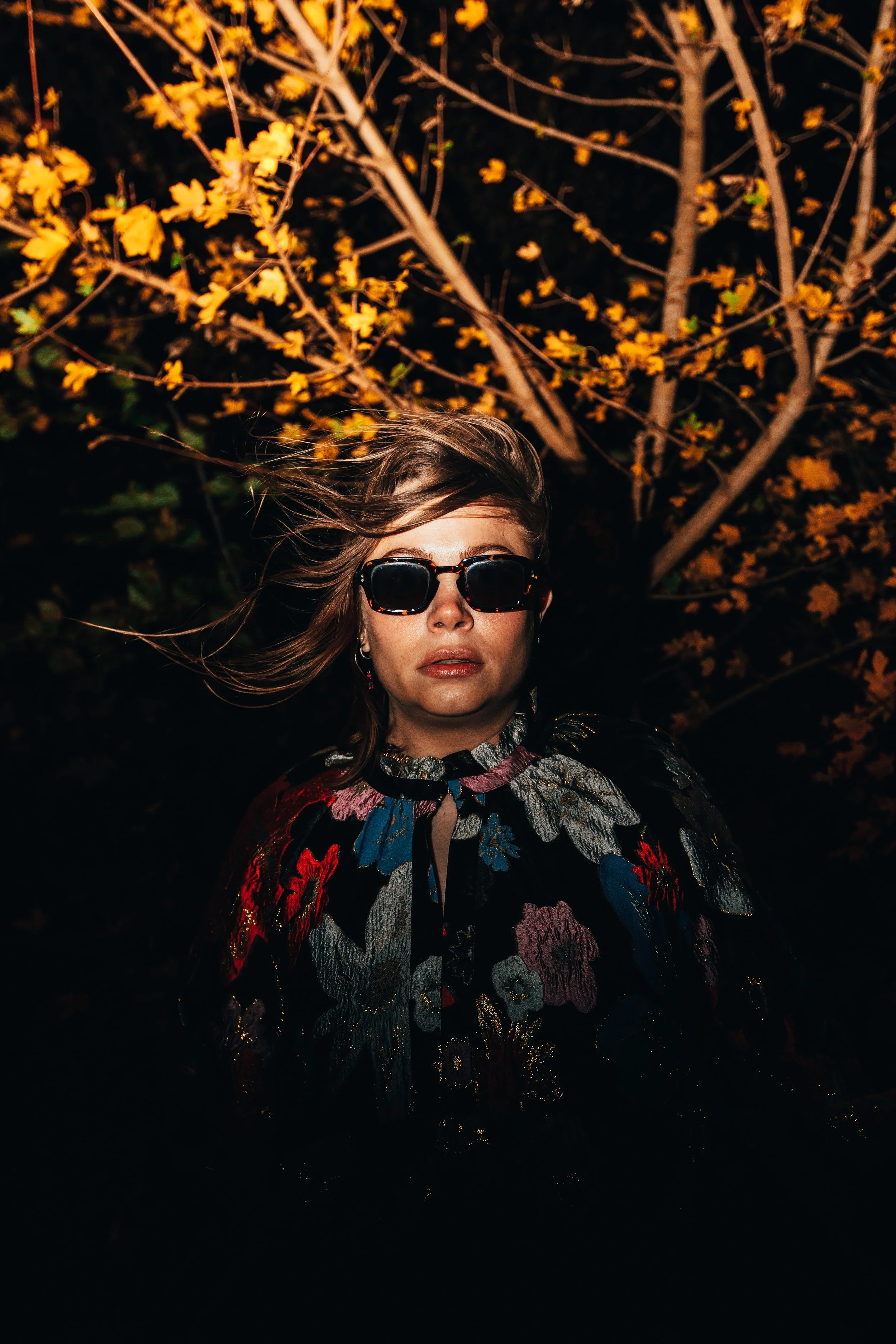 A woman with windblown hair wearing sunglasses and a floral-patterned jacket standing outdoors at night, with a tree with yellow flowers behind her.