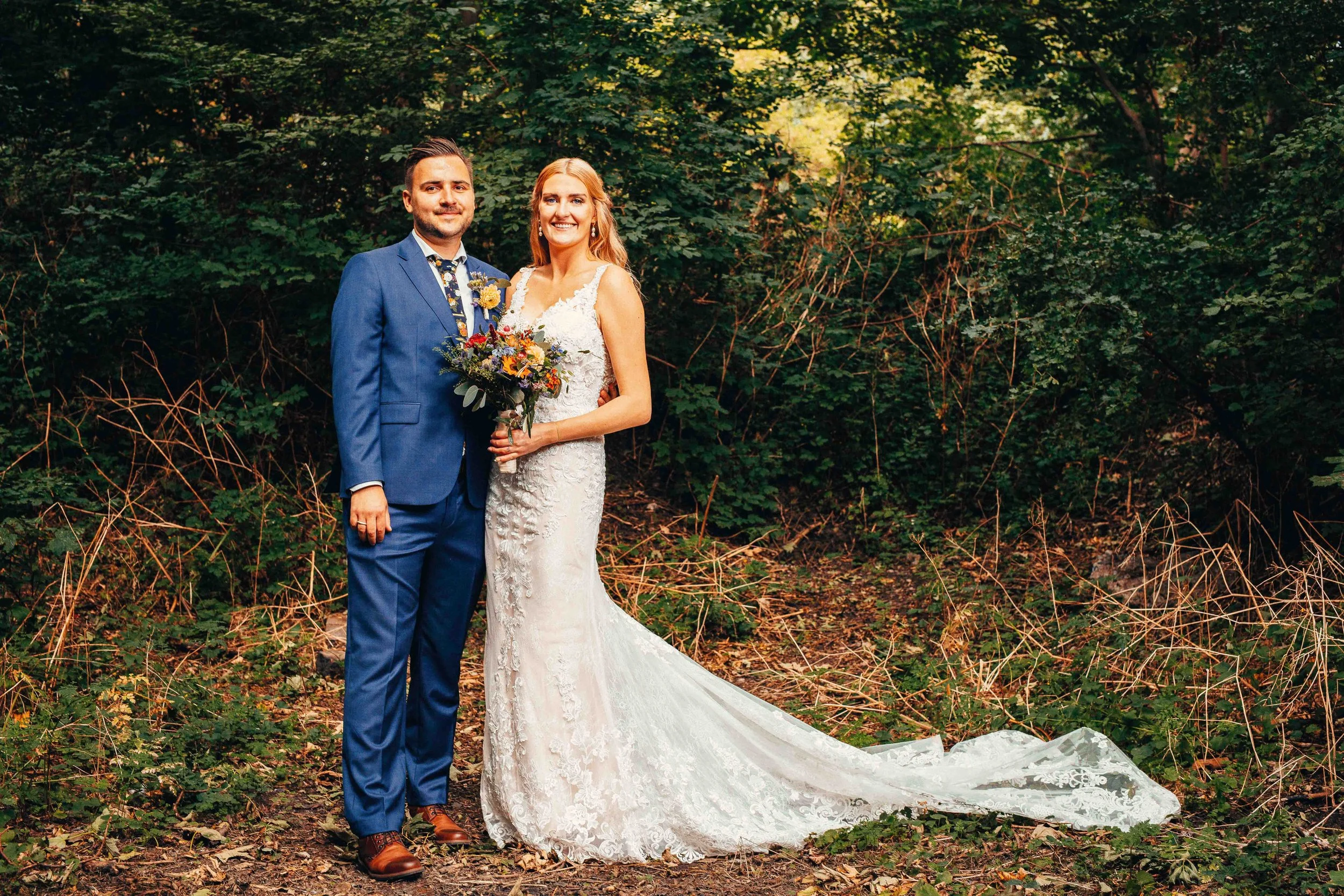 A bride and groom standing together outdoors in a wooded area, smiling at the camera. The bride is wearing a white lace wedding gown and holding a bouquet, while the groom is dressed in a blue suit with a colorful patterned tie.