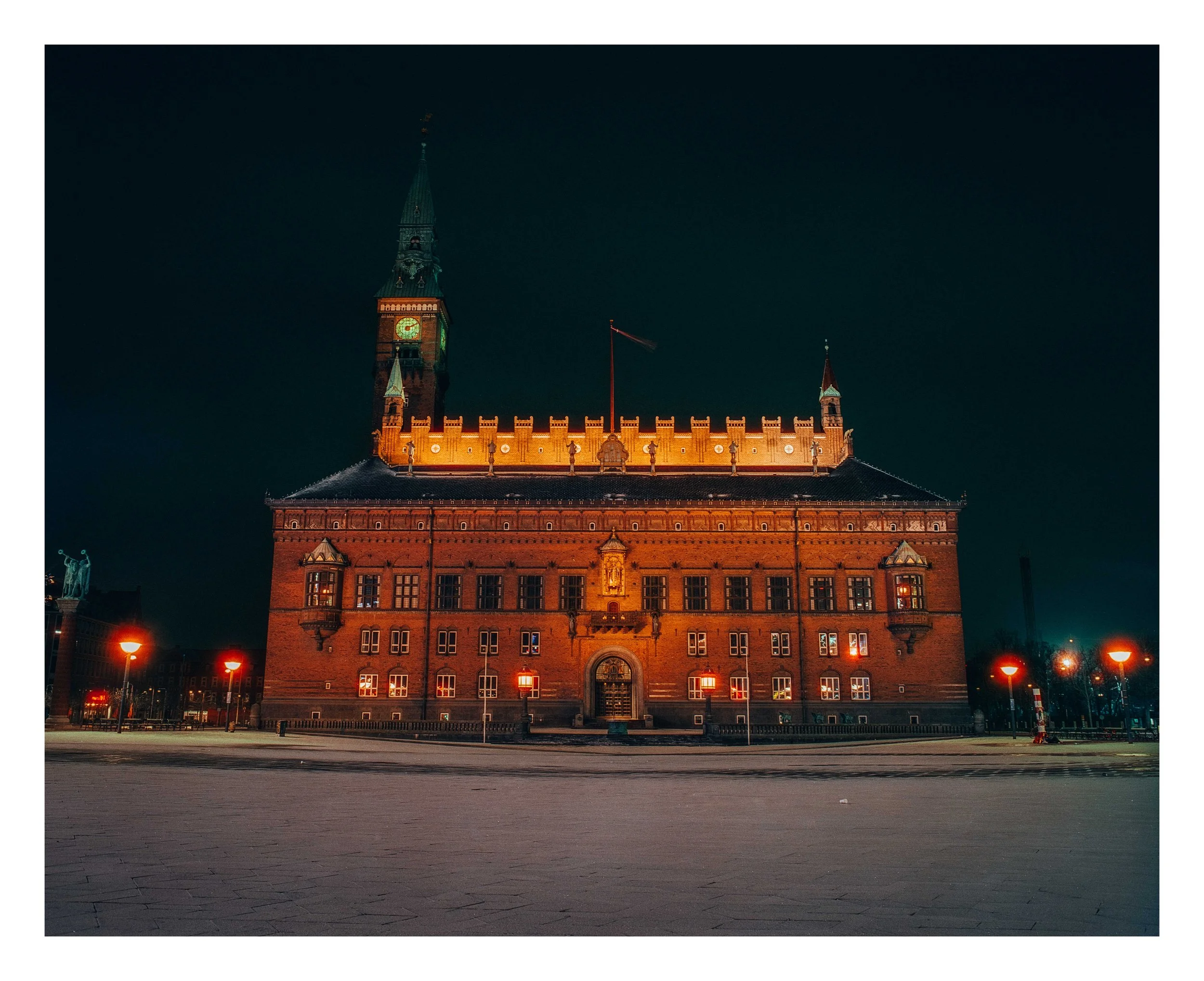 Night view of a historic building with a tall clock tower, illuminated with warm lights against a dark sky, situated in an empty square with street lamps.