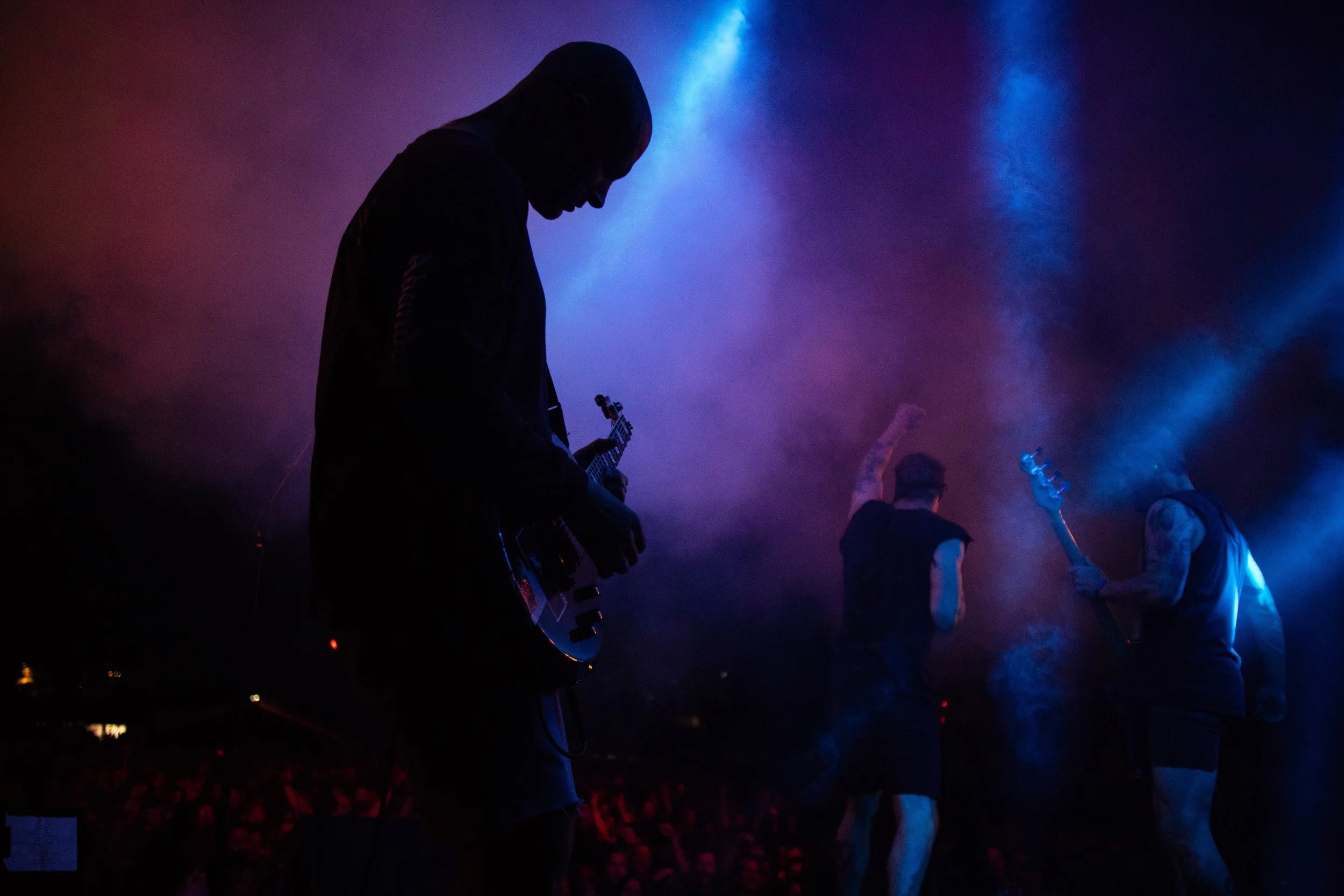 Silhouettes of three people on stage playing musical instruments with colorful stage lighting and smoke effects.