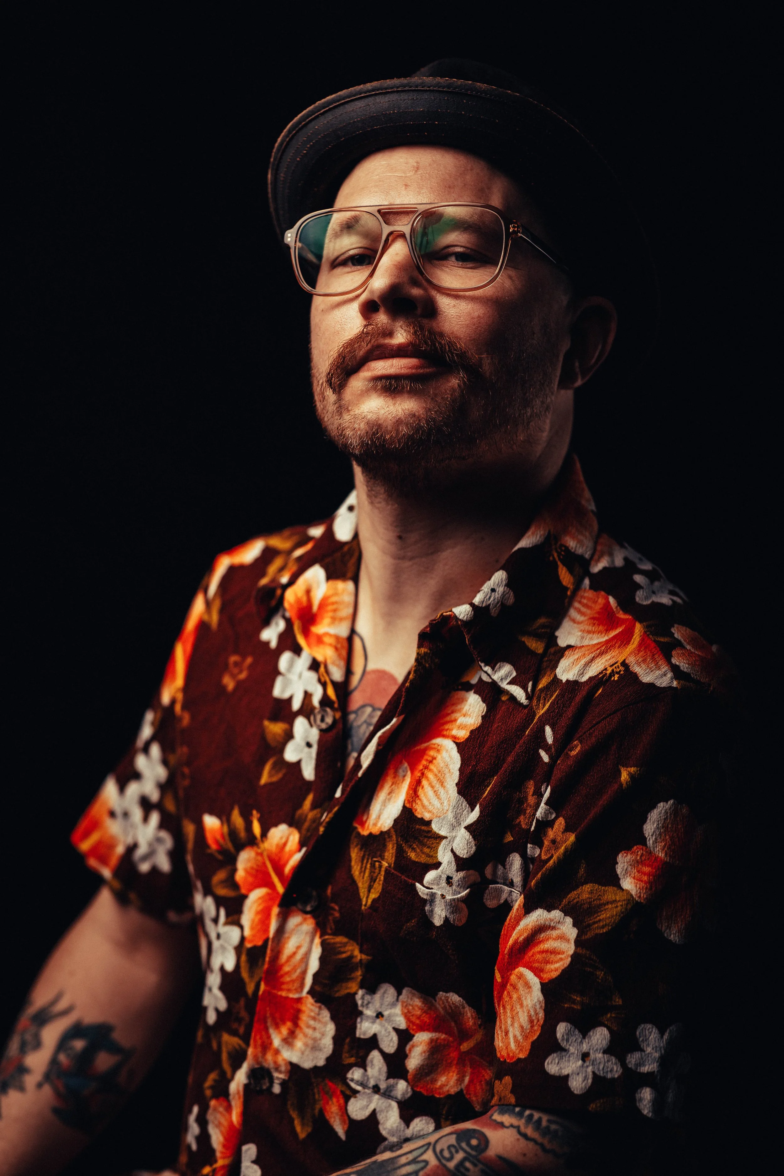 A man with glasses, a mustache, and a beard wearing a floral shirt, a hat, and tattoos on his arm against a dark background.