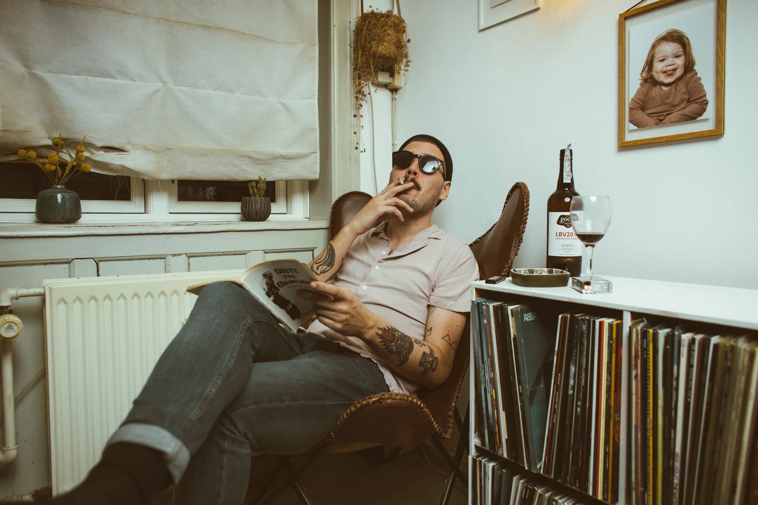 A man with tattoos wearing sunglasses, reading a book, and smoking a cigarette while sitting in a vintage armchair in a cozy room. The room has a window with a blinds and potted plants, a collection of vinyl records on a shelf, a framed photo of a sm