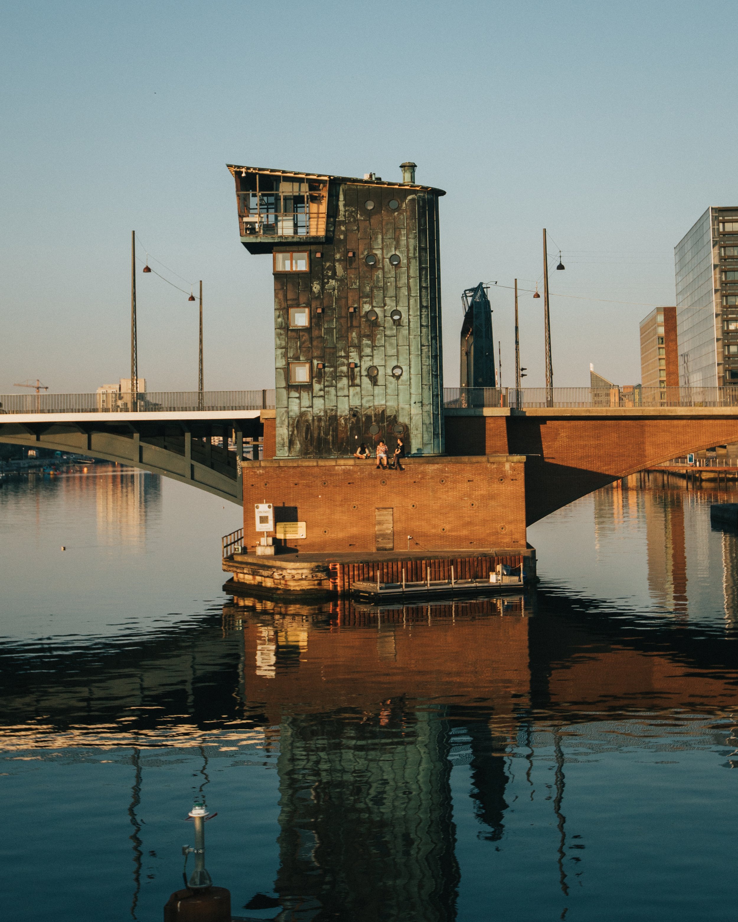 A unique modern building with an irregular shape, situated on a pier by the water, with nearby buildings and a bridge in the background during sunset.