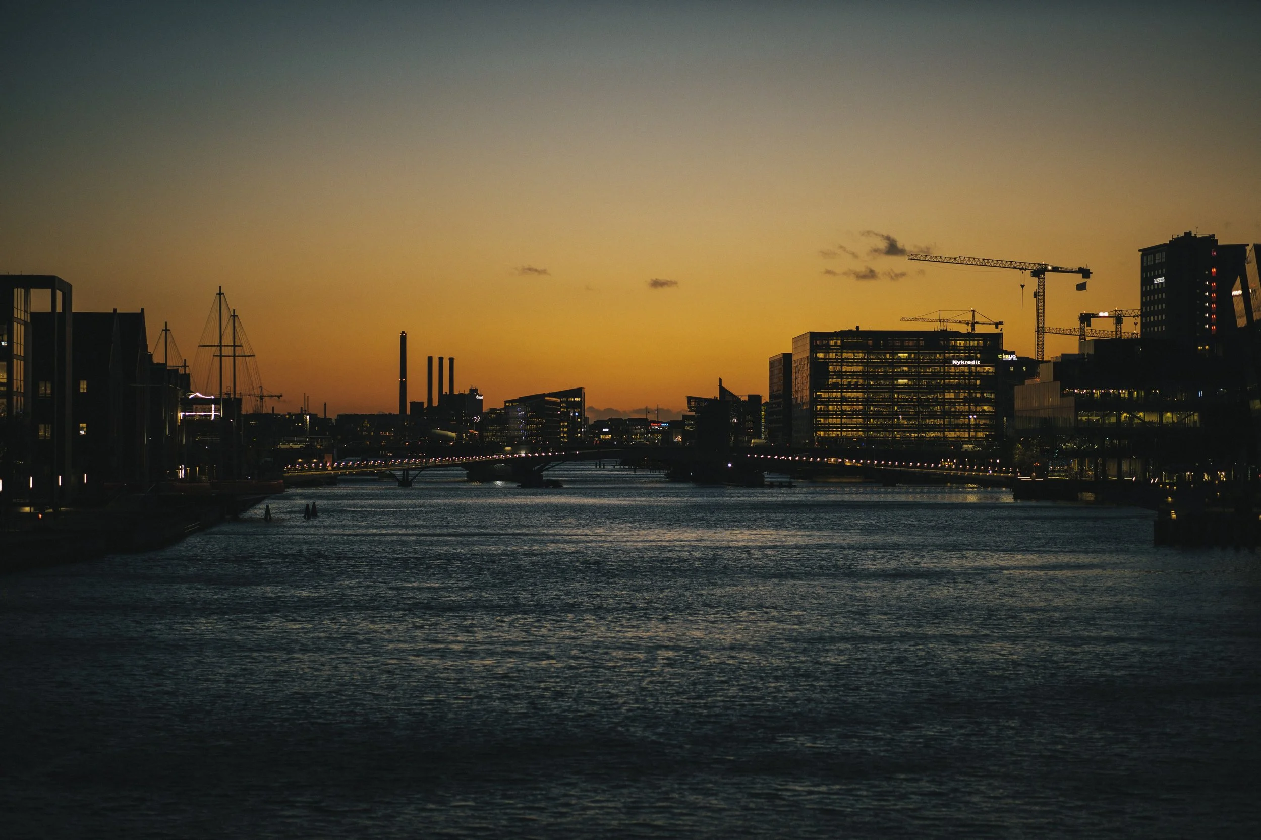 Sunset over a city skyline with water in the foreground, multiple buildings including glass offices, construction cranes, and some boats on the water.